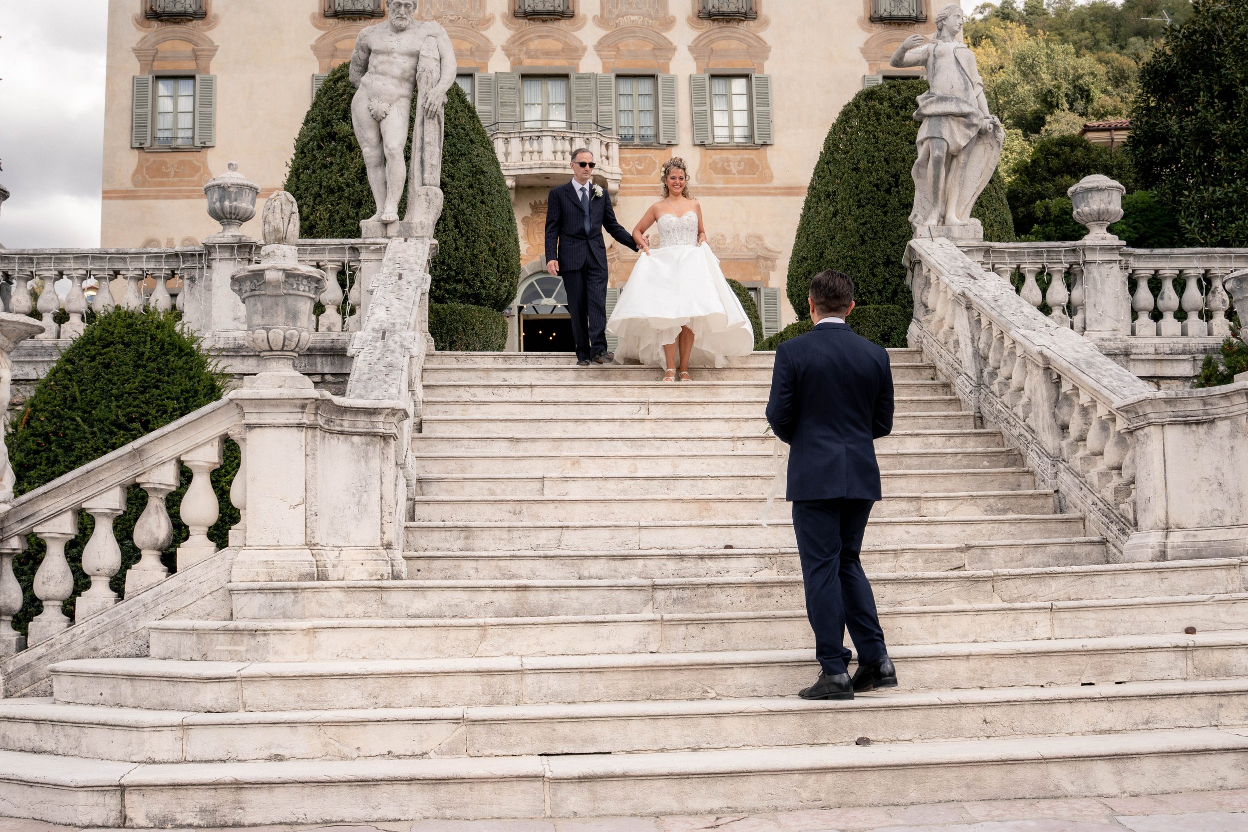 Bride and groom walking down stone stairs outside a historic building with statues and greenery, wedding celebration.