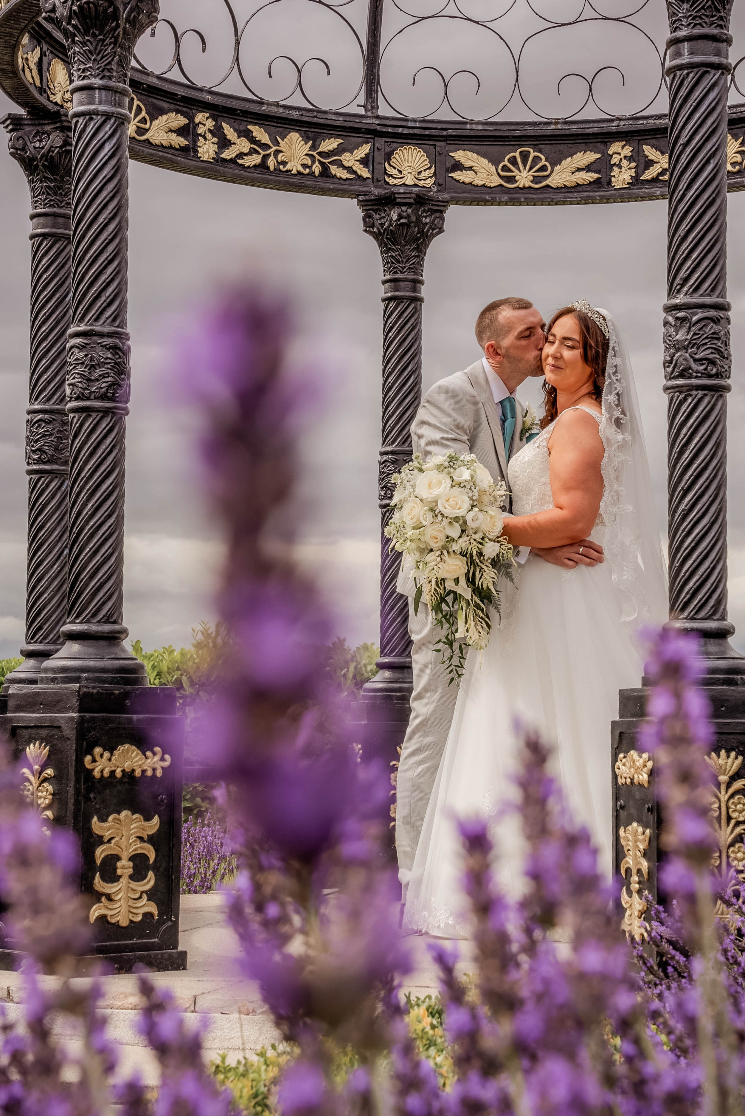 A bride and groom share a kiss under an ornate black and gold gazebo, with purple flowers in the foreground.
