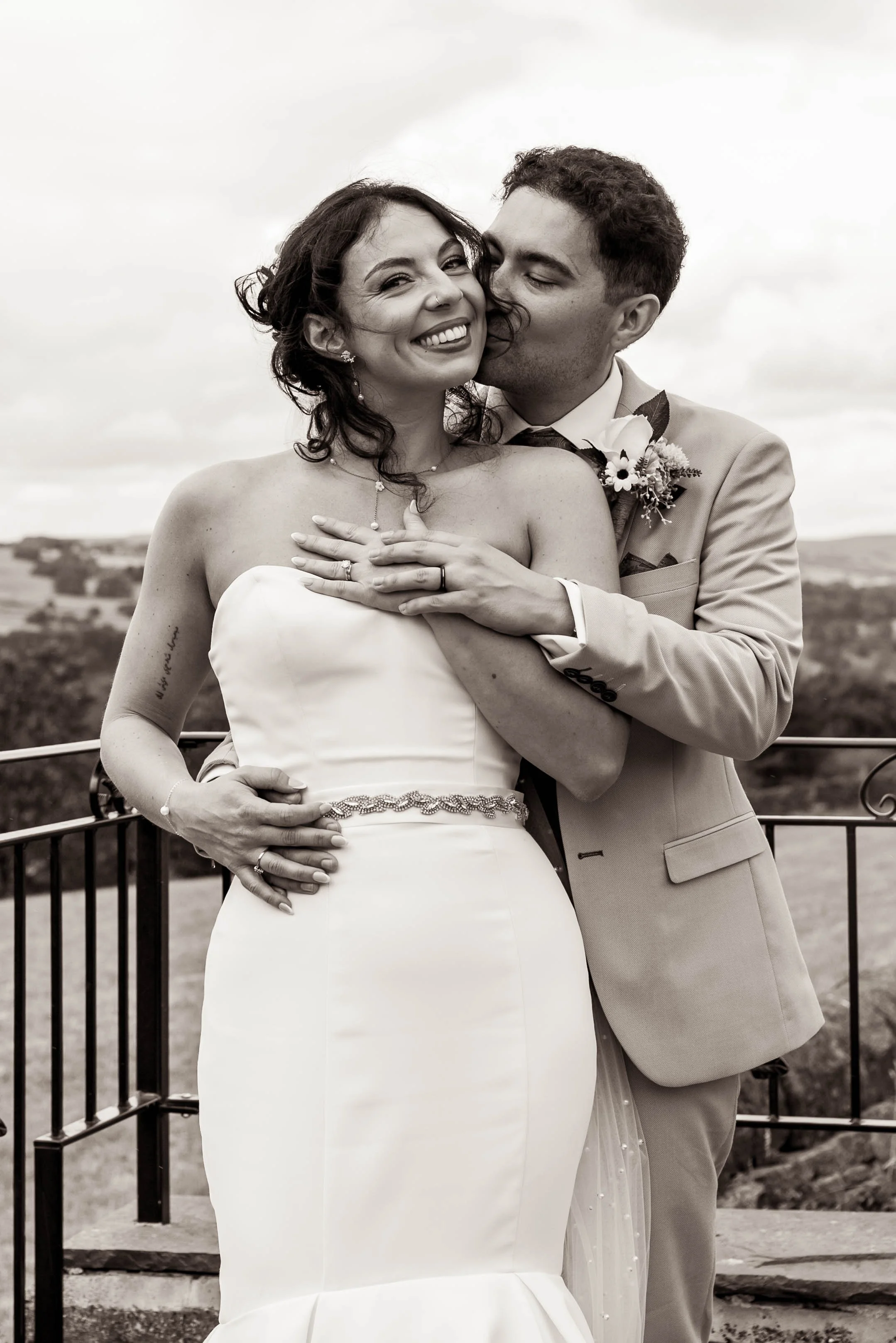 A black and white photo of a bride and groom smiling and embracing outdoors on her wedding day, with rolling hills in the background.