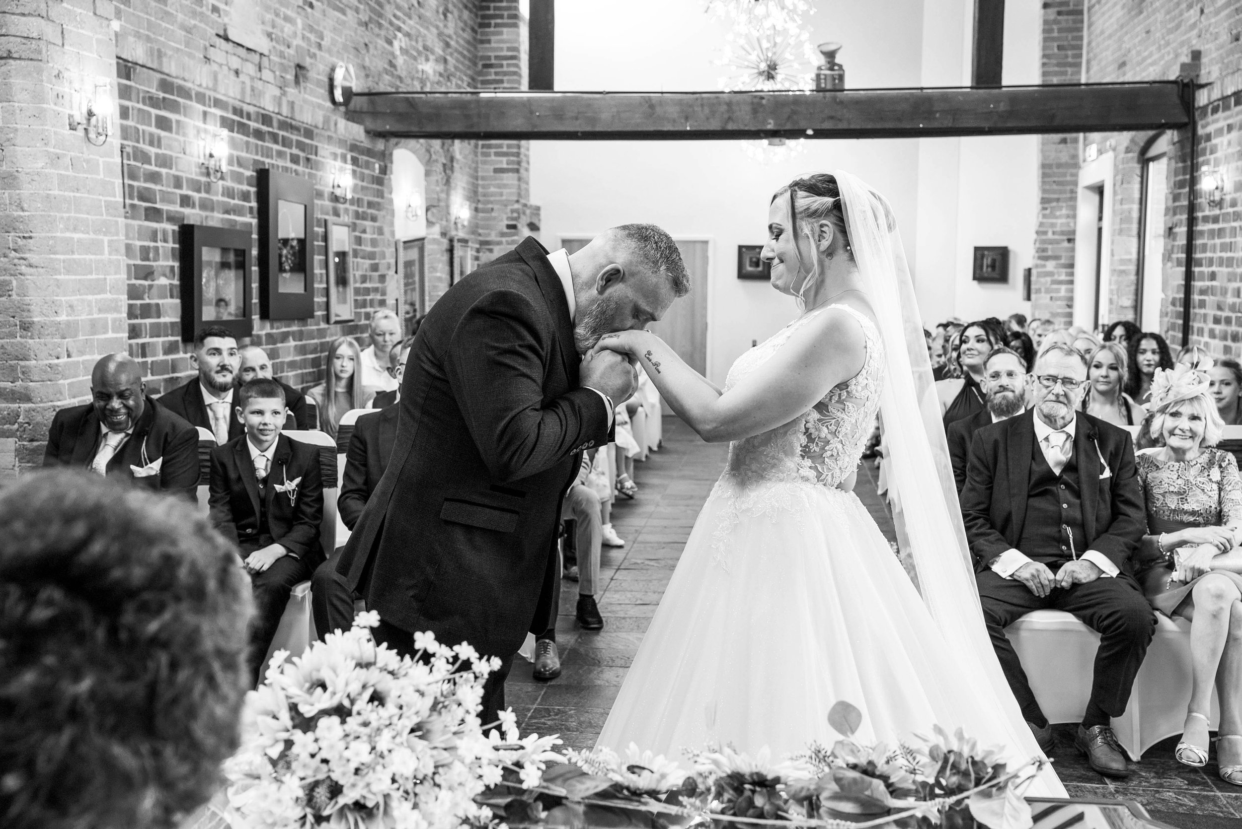 A black-and-white photo of a wedding ceremony with the groom kissing the bride's hand. The bride is wearing a white lace wedding dress with a veil, and the groom is in a dark suit. The ceremony is taking place indoors with brick walls, and guests are