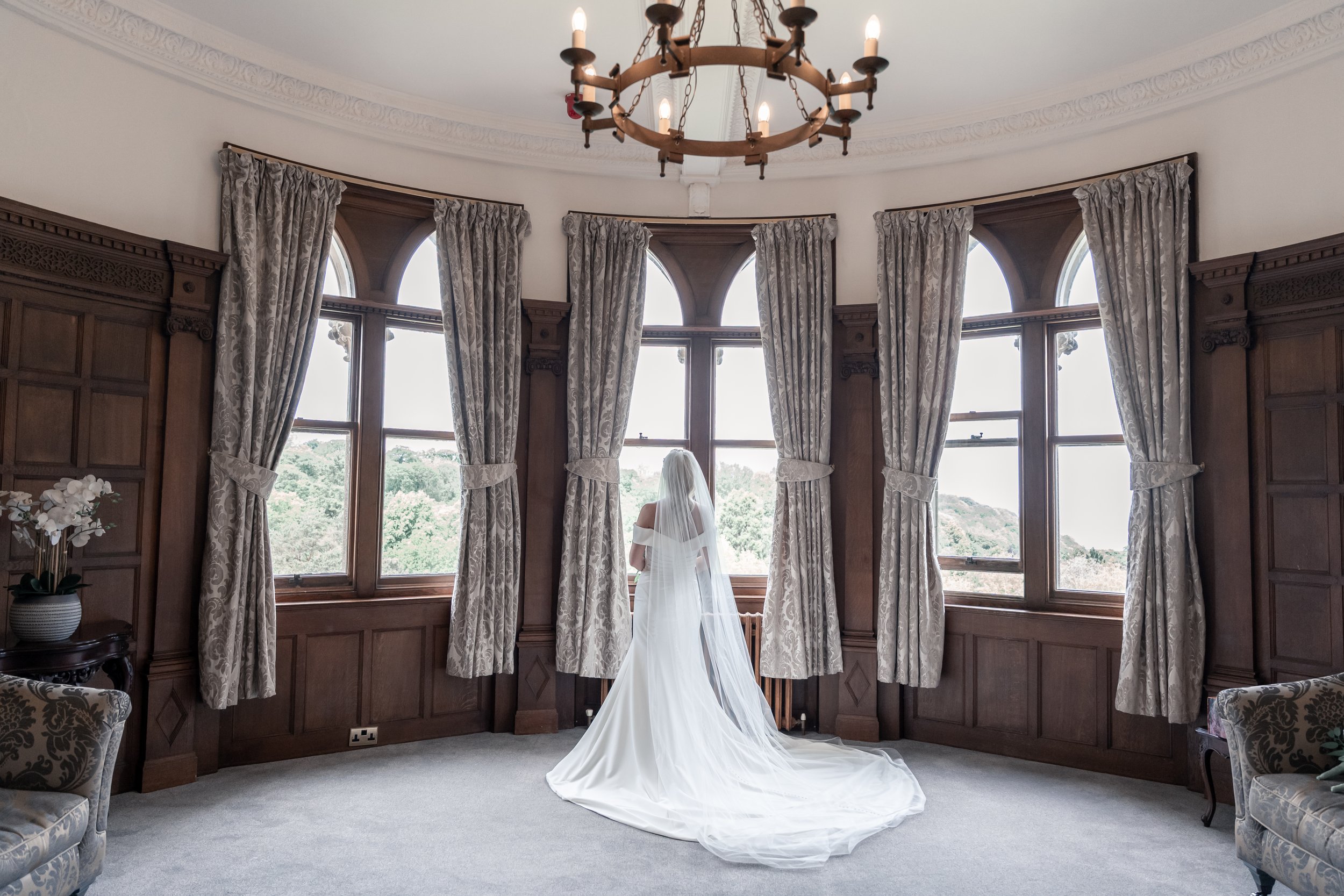 A bride in a white wedding dress and veil standing in front of a large window with beige curtains in a wood-paneled room, with a chandelier hanging from the ceiling.
