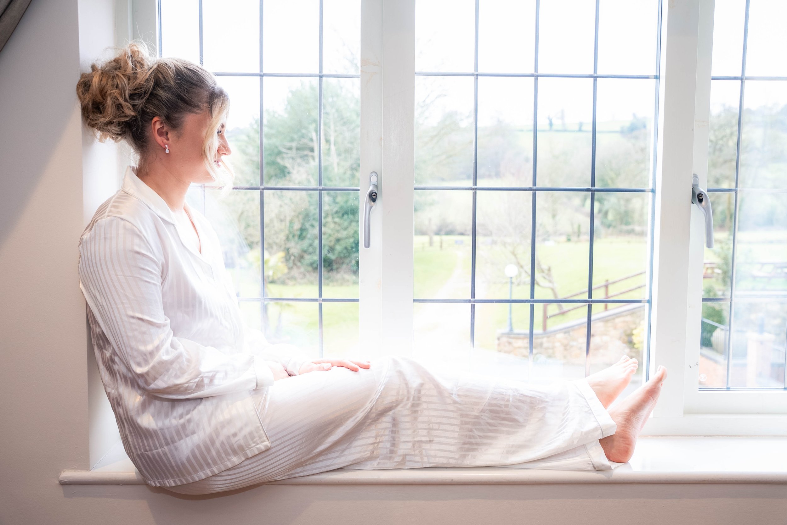 Woman in pajamas sitting on a window sill, looking outside on a bright day.
