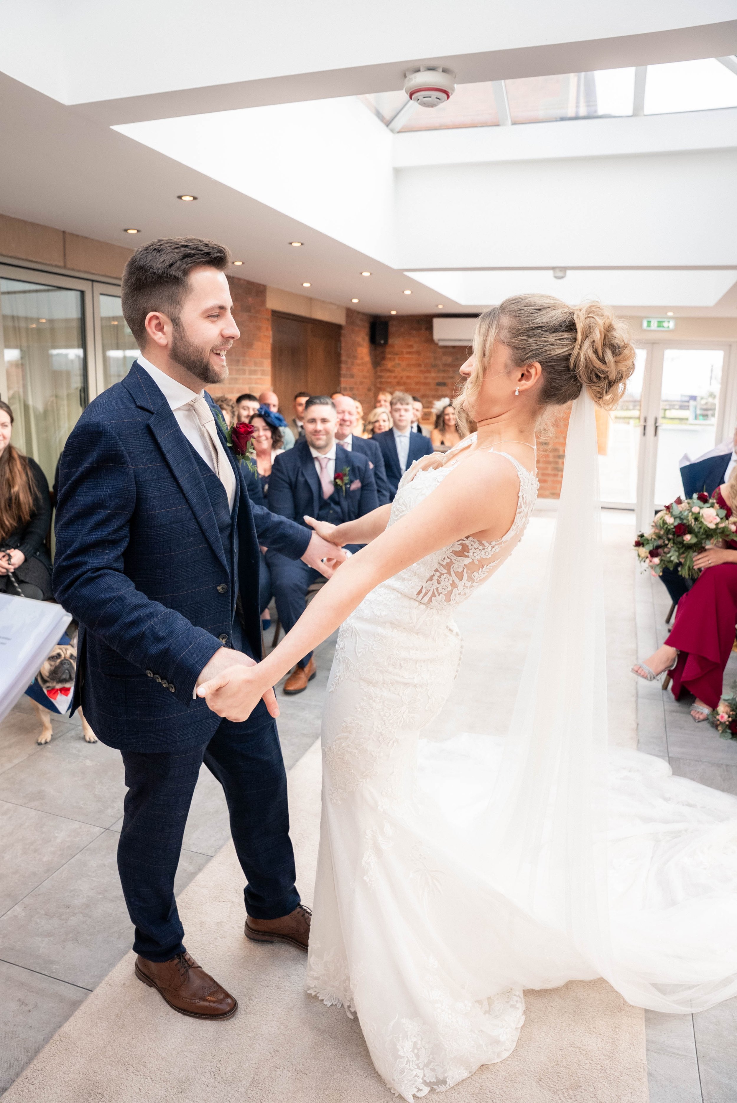Bride and groom holding hands and smiling during their wedding ceremony indoors with seated guests in the background.