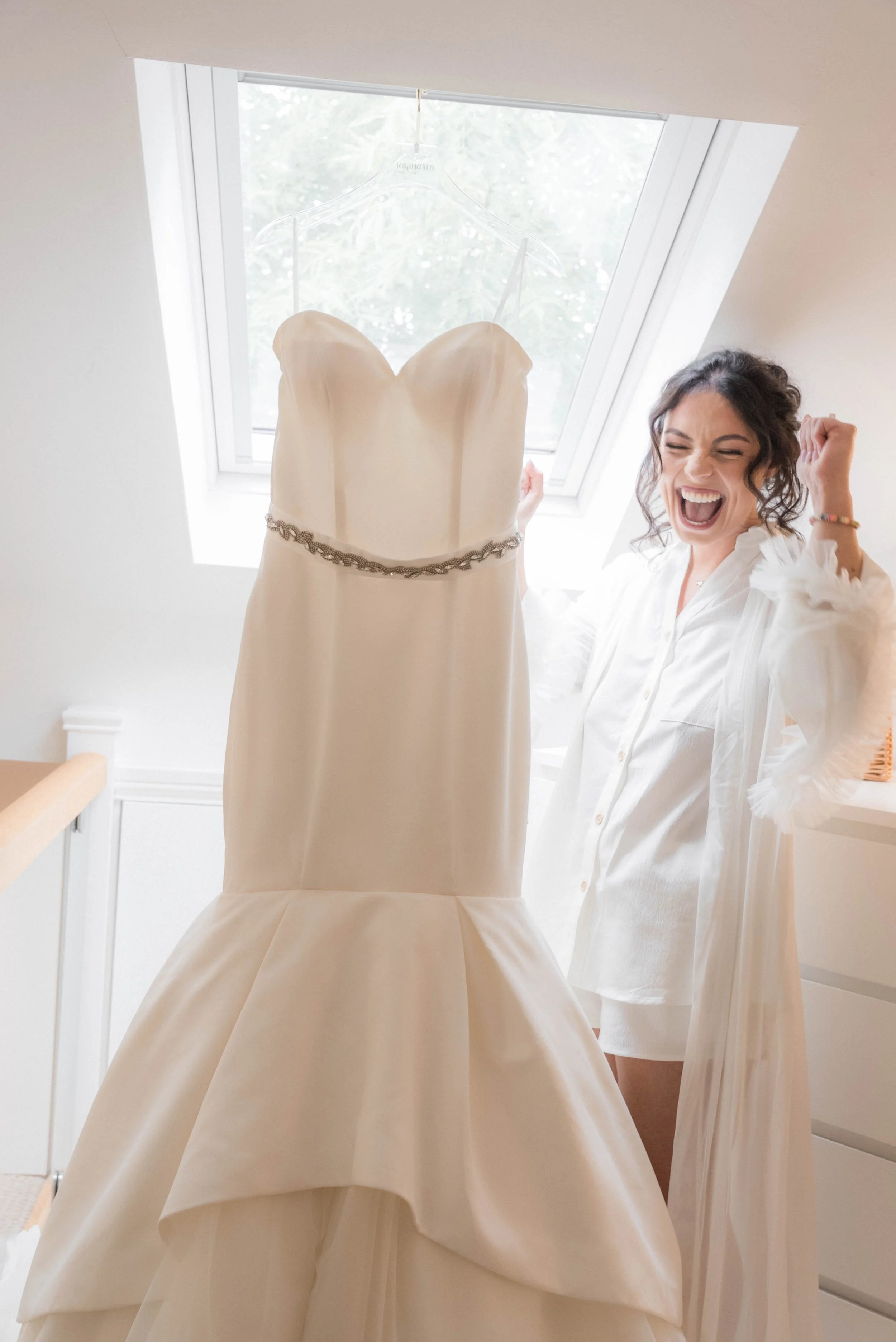 Woman celebrating with a wedding dress hanging on a hanger in front of a window.