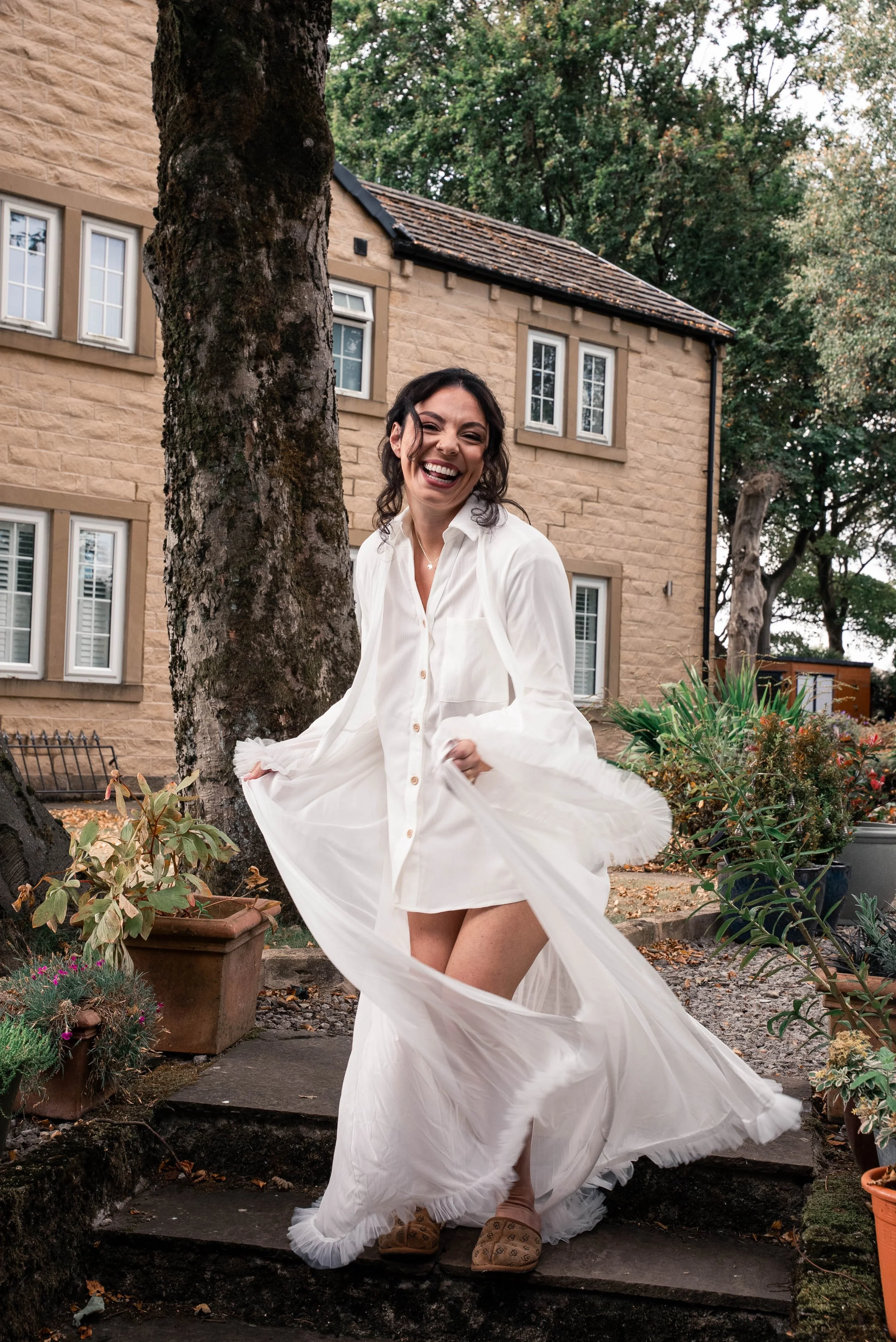 A woman in a white dress is smiling and dancing outdoors near a large tree, with a stone house and plants in the background.