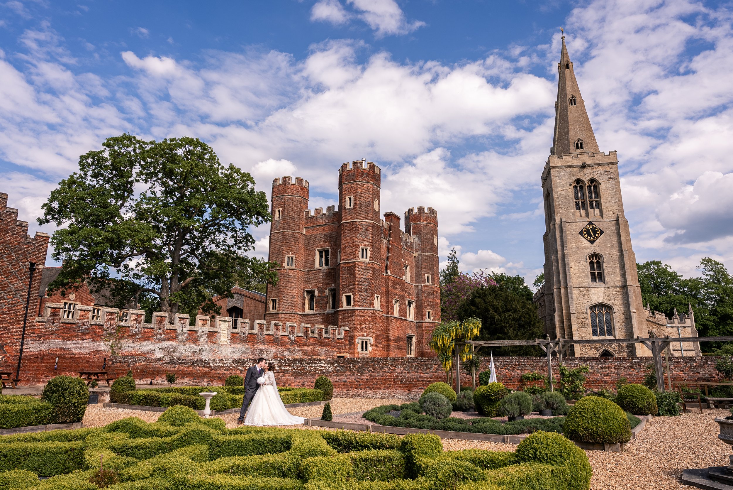 A bride and groom in wedding attire standing in a garden in front of a historic castle with a tall church steeple, brick walls, and a blue sky with clouds.