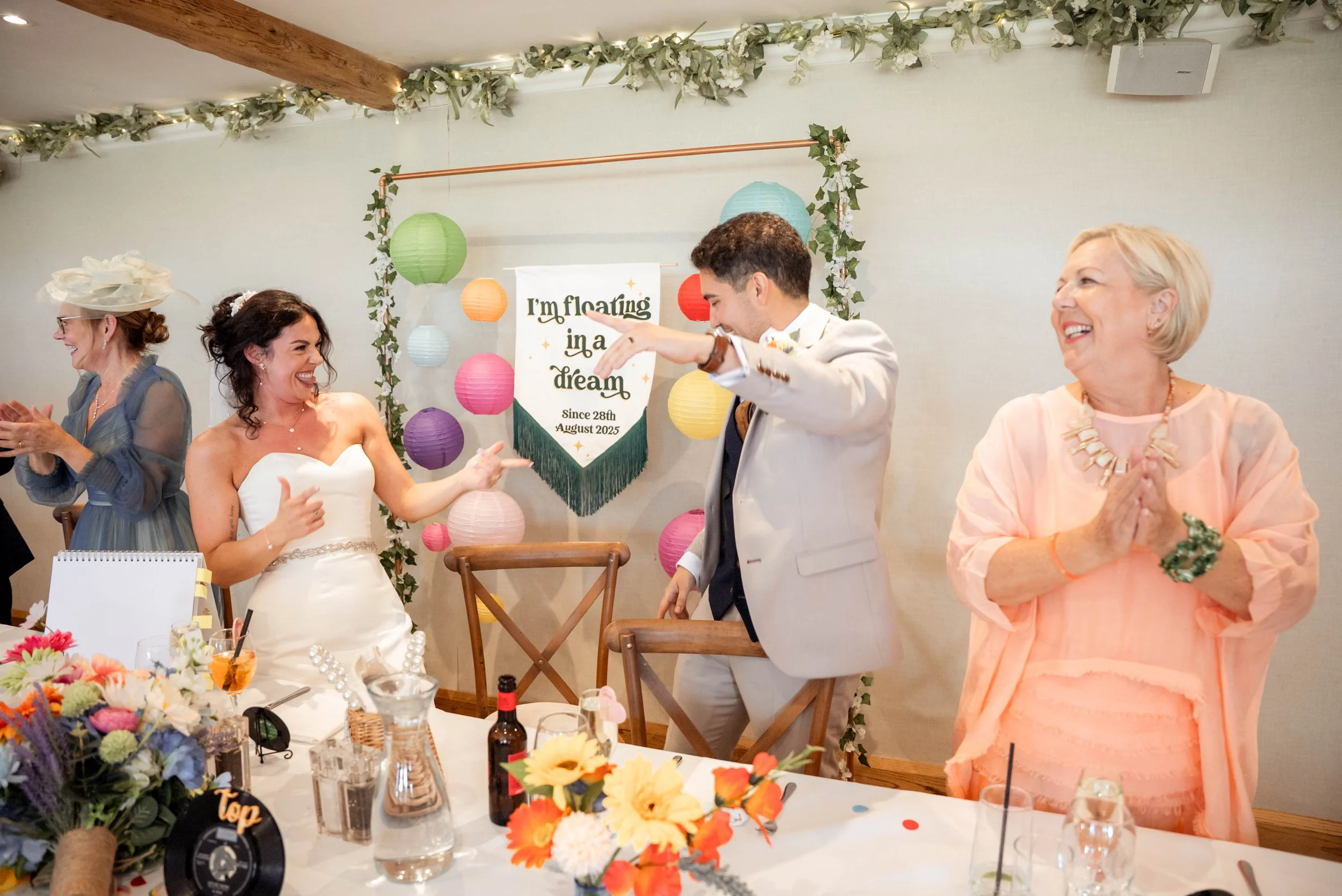 A wedding celebration with a bride in a white strapless gown and a groom in a light-colored suit, both smiling and looking at each other. An older woman on the right is clapping and smiling, while a woman in a blue dress and hat is on the left clappi