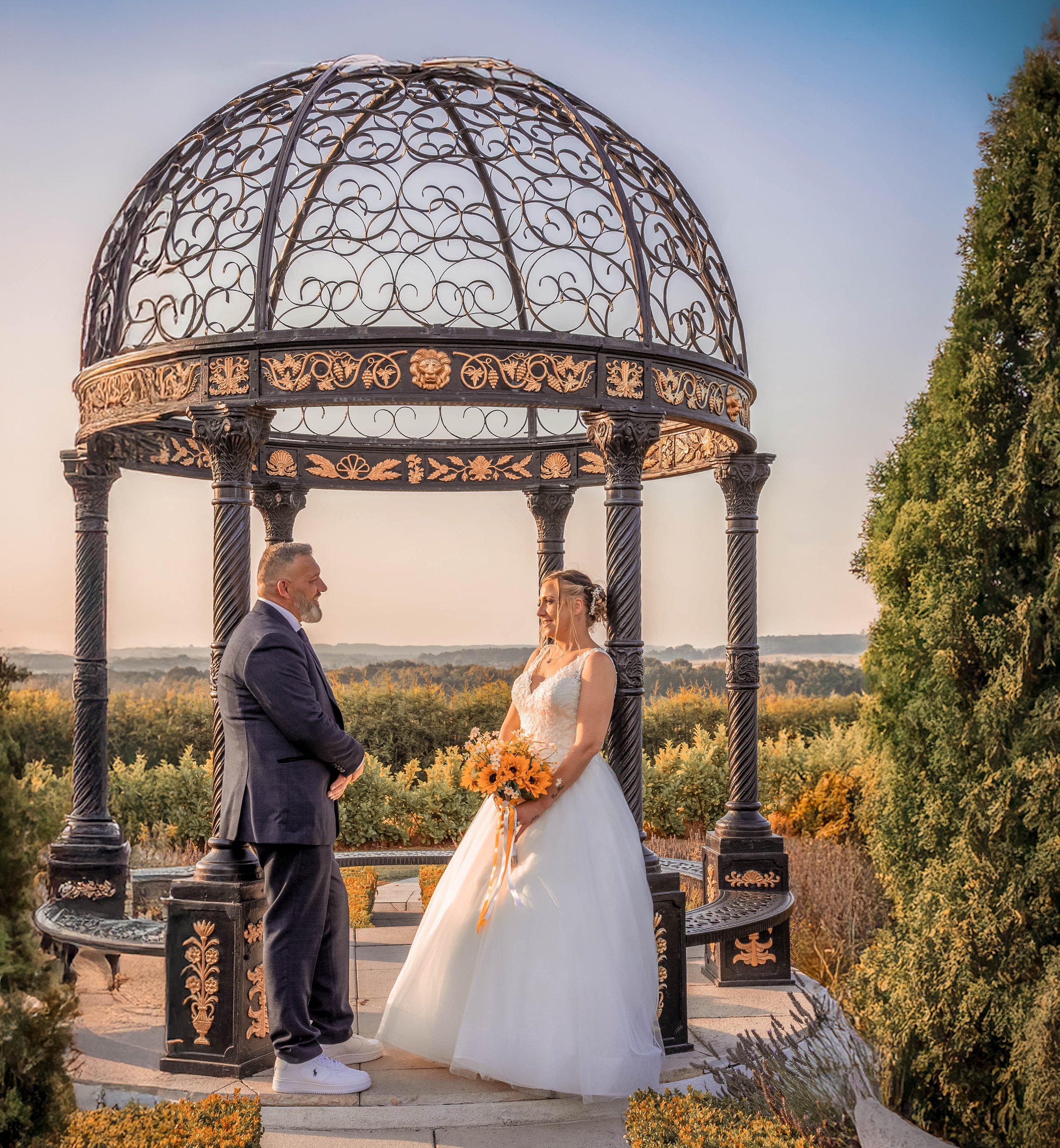 A couple dressed in wedding attire standing under an ornate, black and gold wrought iron gazebo with a scenic outdoor landscape in the background.