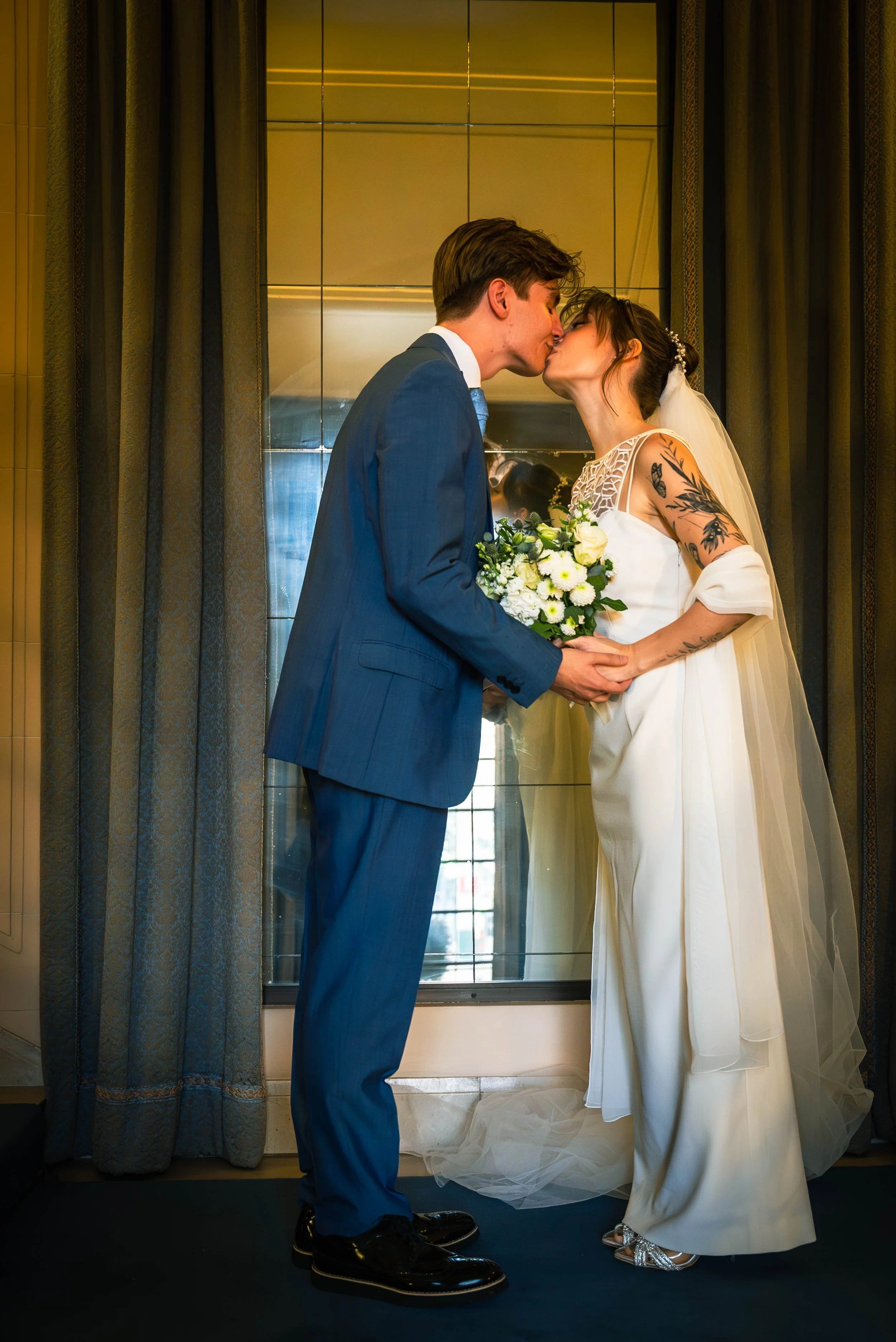 A couple dressed in wedding attire sharing a kiss, holding a bouquet of white flowers, standing in front of a mirror with curtains on either side.