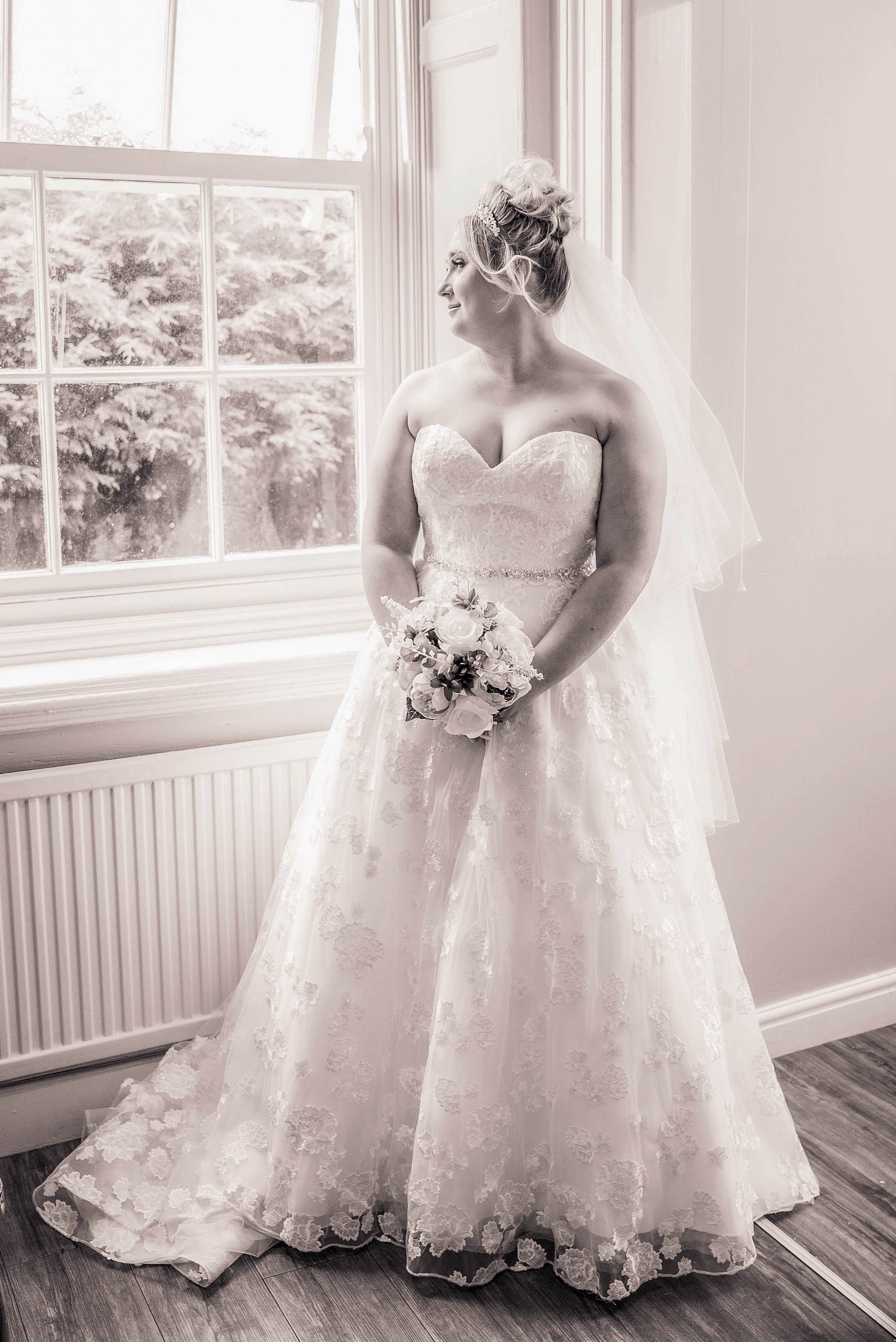 A bride in a strapless wedding gown holding a bouquet, standing by a window with natural light, looking to the side.