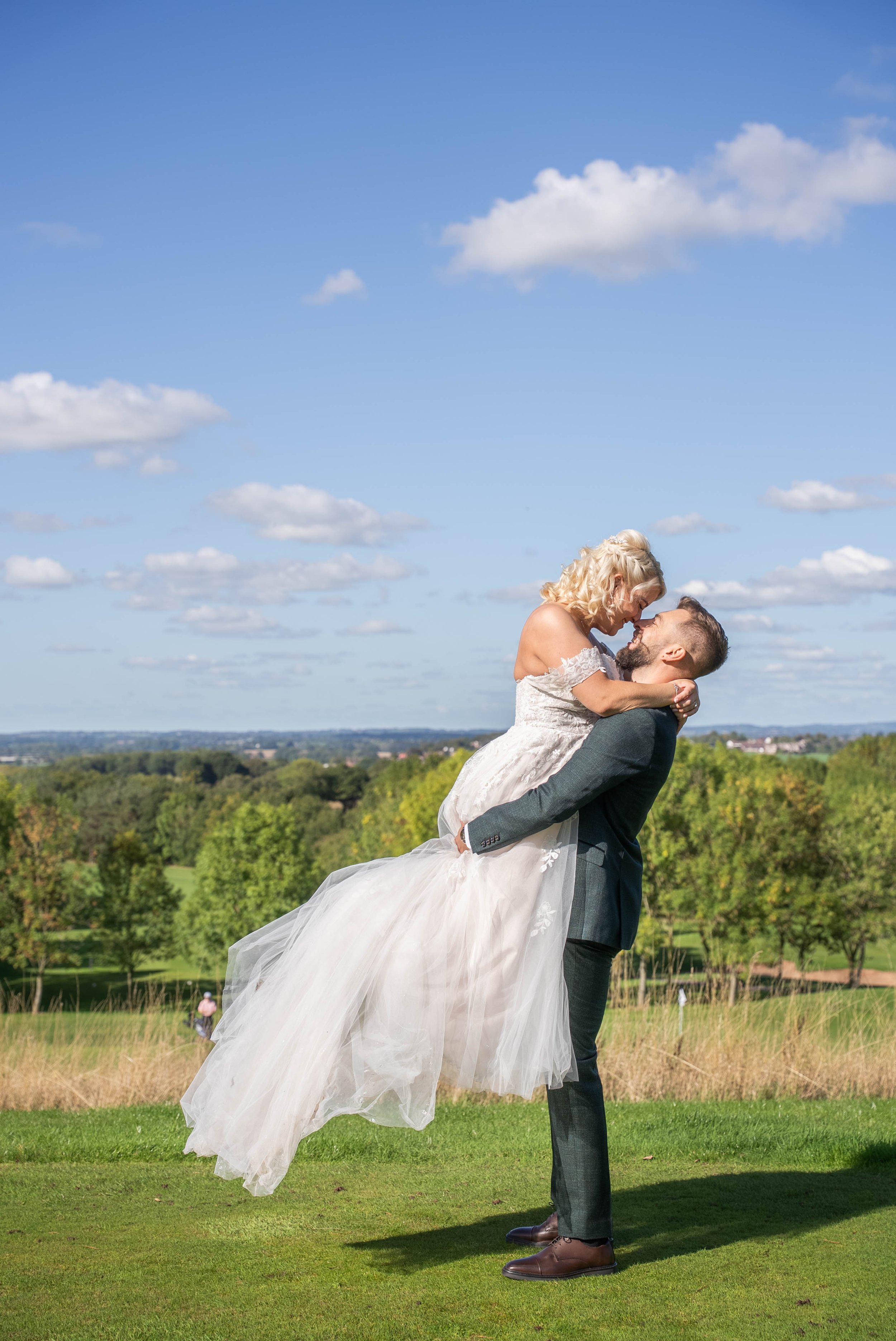 A man in a suit lifting a woman in a wedding dress on a grassy hill, with trees and a blue sky with scattered clouds in the background.