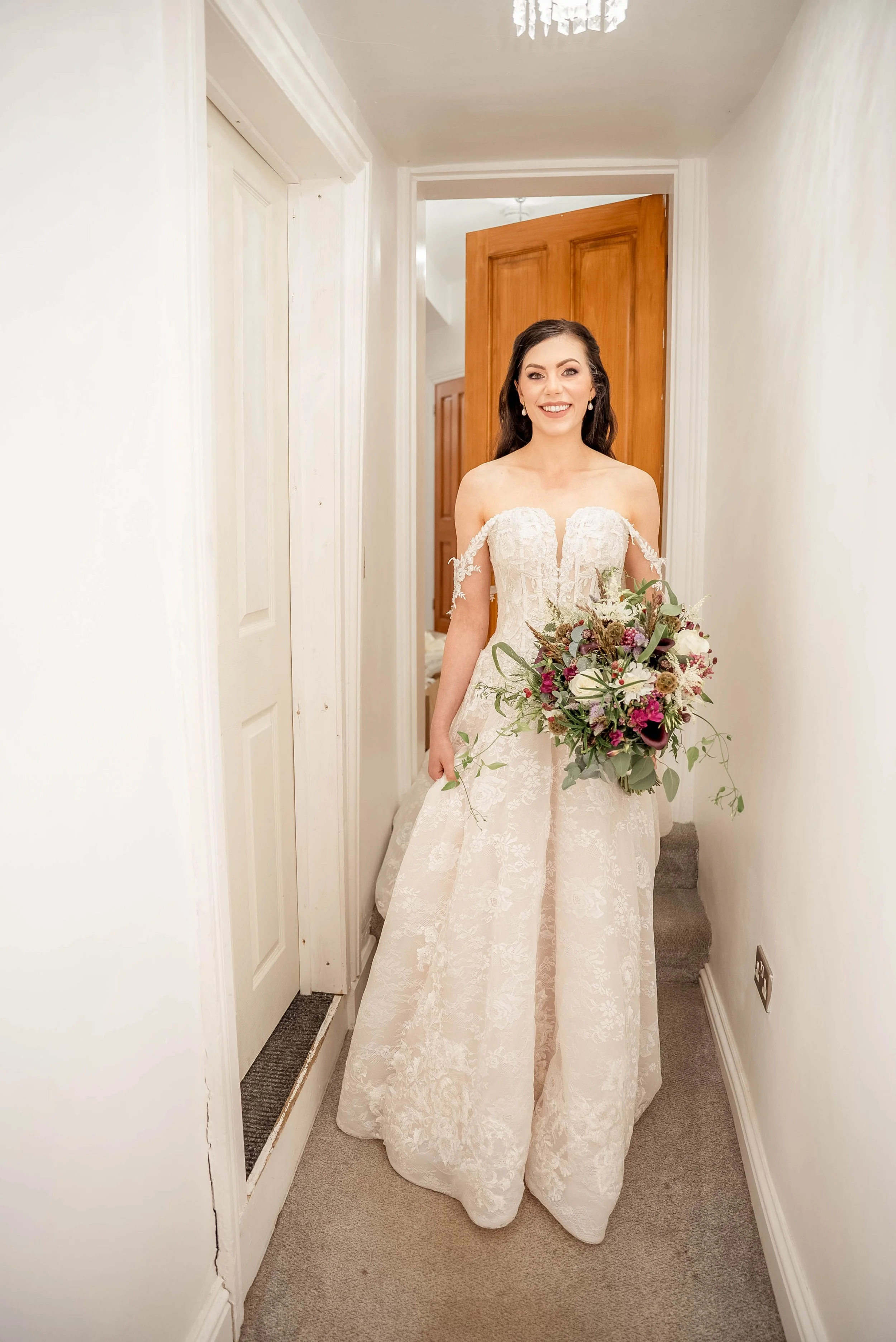 A bride in a white wedding gown holding a bouquet, standing in a hallway with white walls and a wooden door.