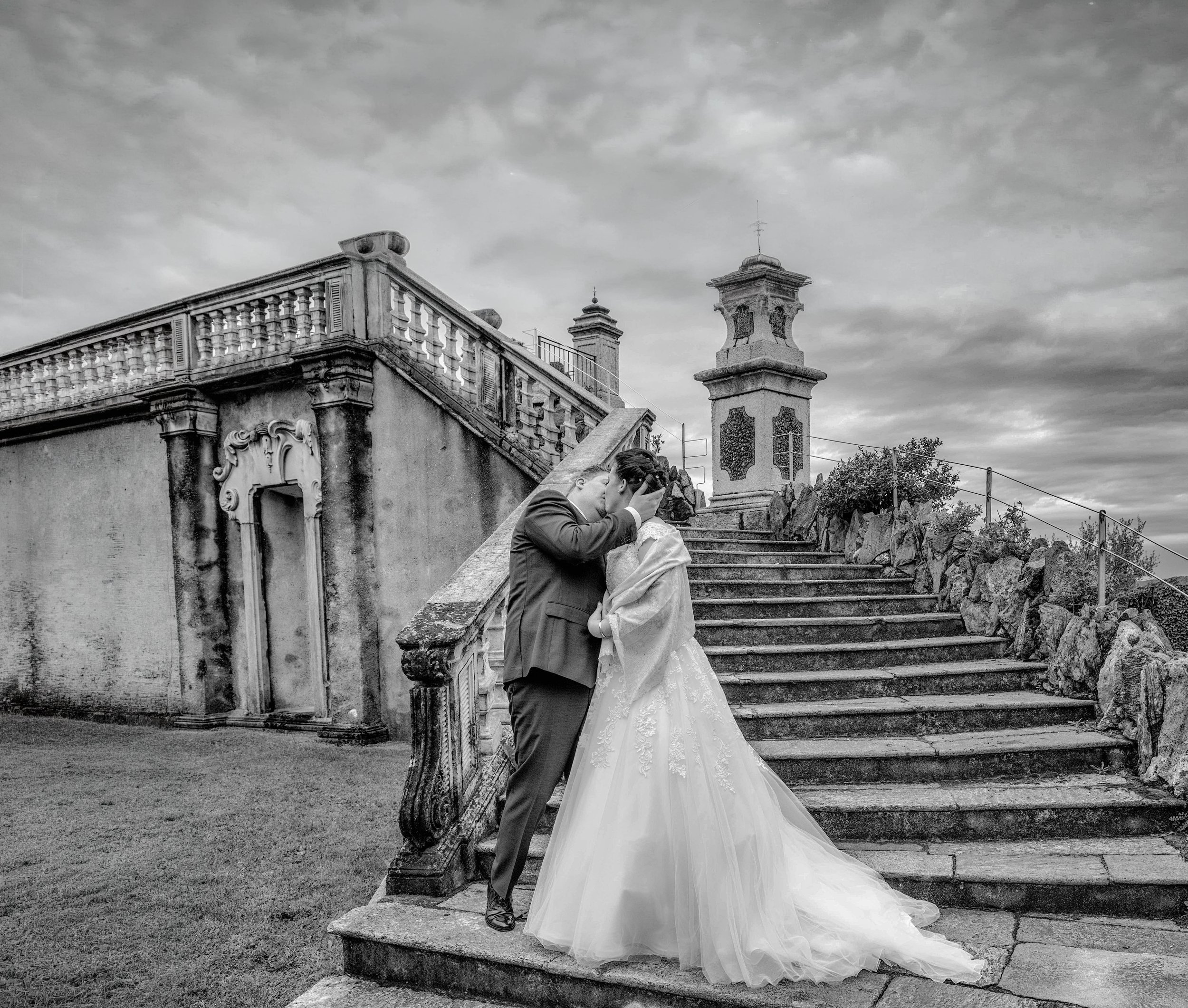 A black and white photo of a bride and groom kissing on stone steps outdoors, with a decorative stone pillar and a building railing visible behind them. The bride wears a white wedding gown with lace details, and the groom is dressed in a suit. The s