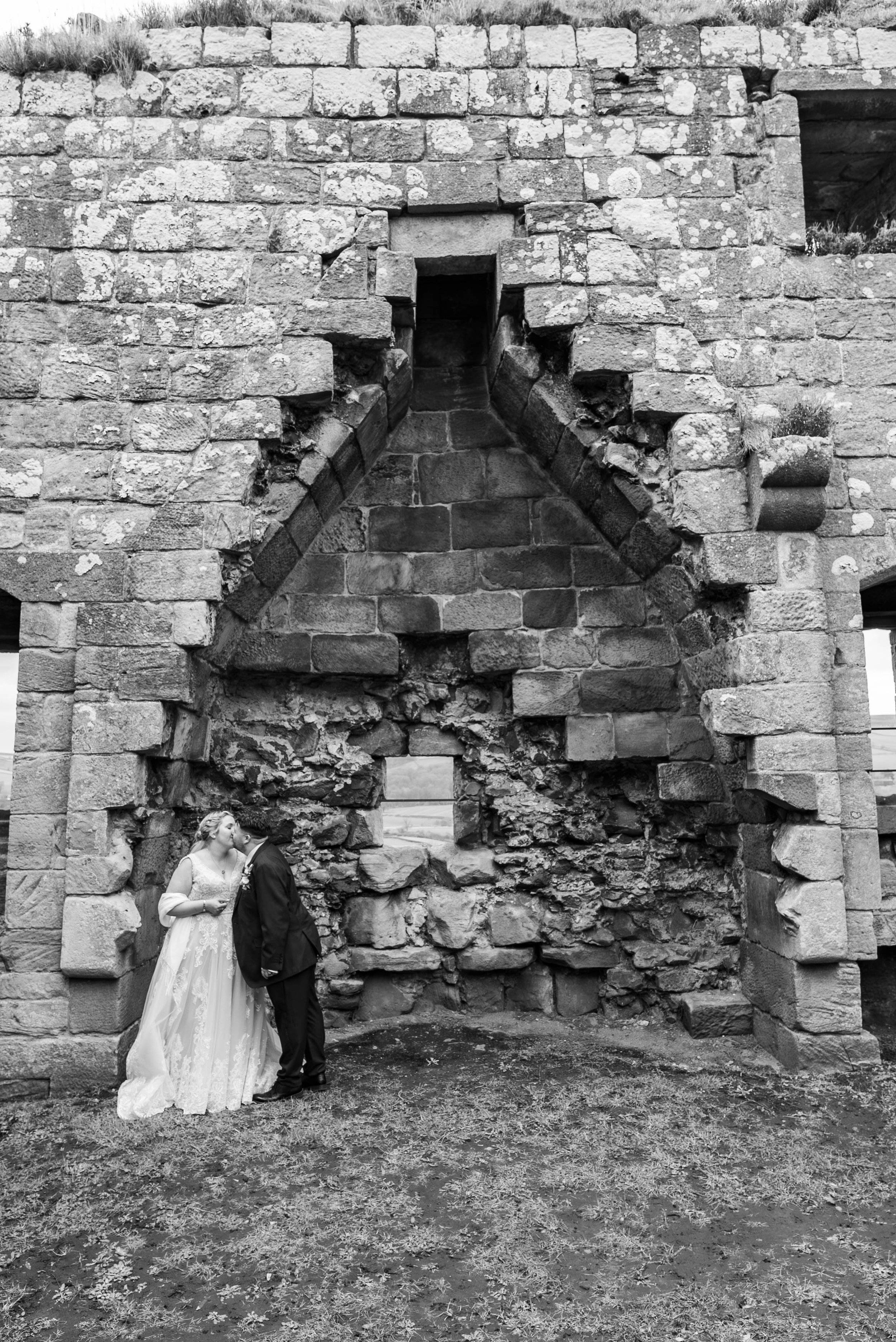A wedding couple standing close together in front of a large, ancient stone wall with an archway. The bride is in a white wedding gown, and the groom is in a dark suit. They are touching foreheads and appear to be sharing a tender moment.