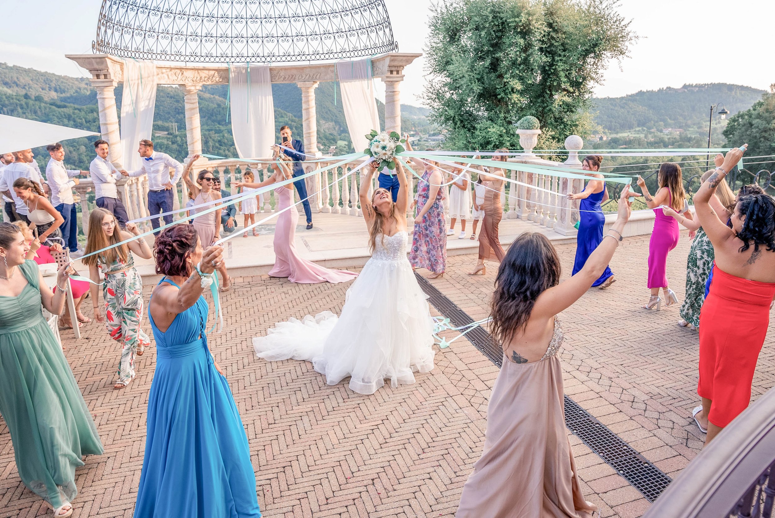 A bride in a white gown celebrating with guests at her wedding reception outdoors on a patio with scenic hills in the background. Guests are dancing and holding ribbons radiating from the bride while a man in a suit takes a photo.