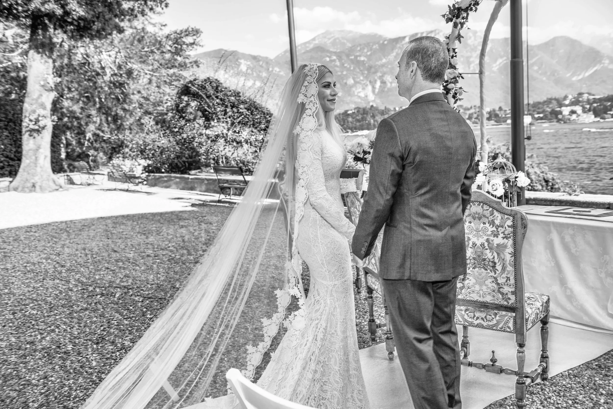 A bride and groom exchanging vows at a lakeside outdoor wedding ceremony, with mountains in the background.