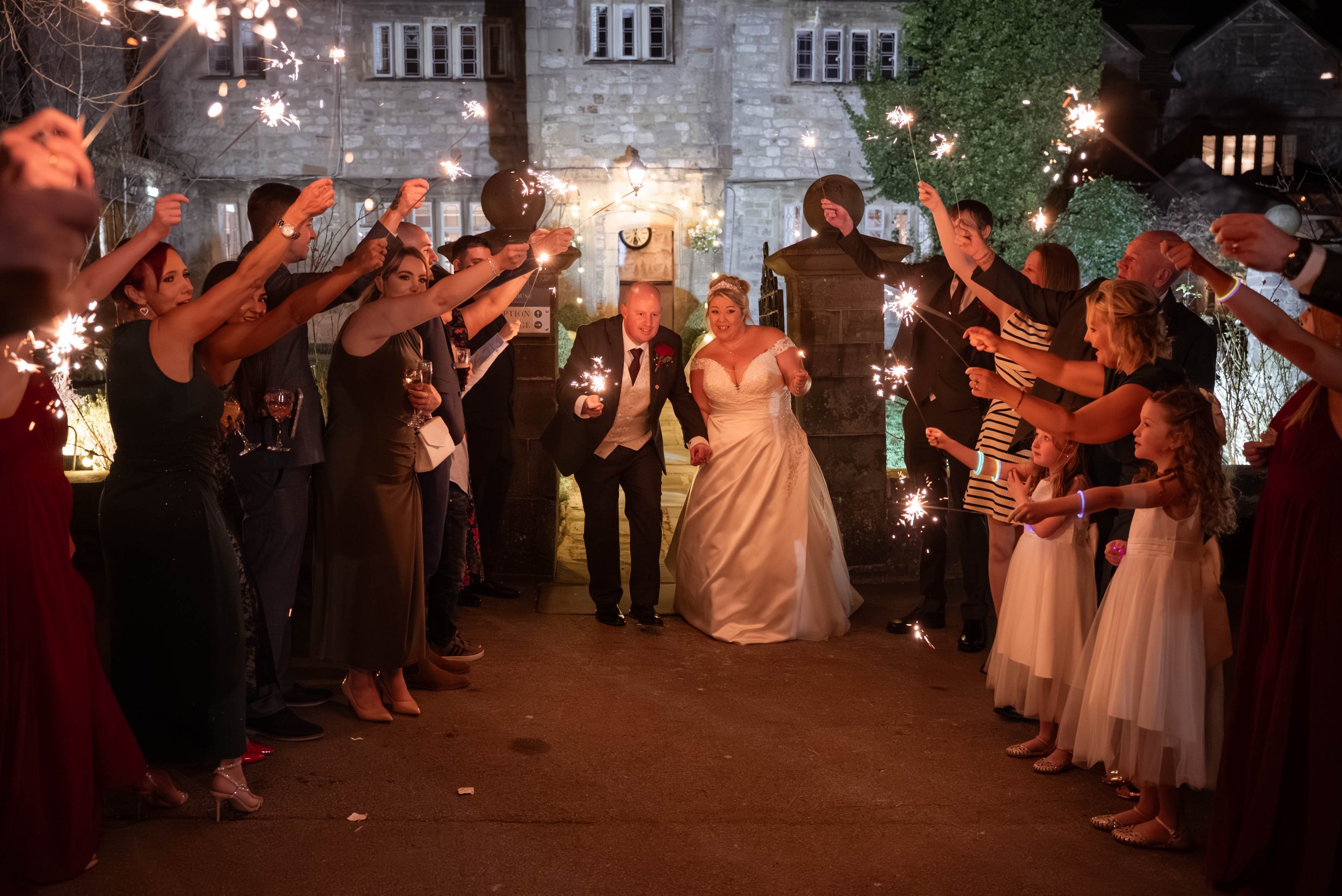 Bride and groom walking through a sparkler tunnel during their wedding celebration outside at night with guests on both sides holding sparklers.