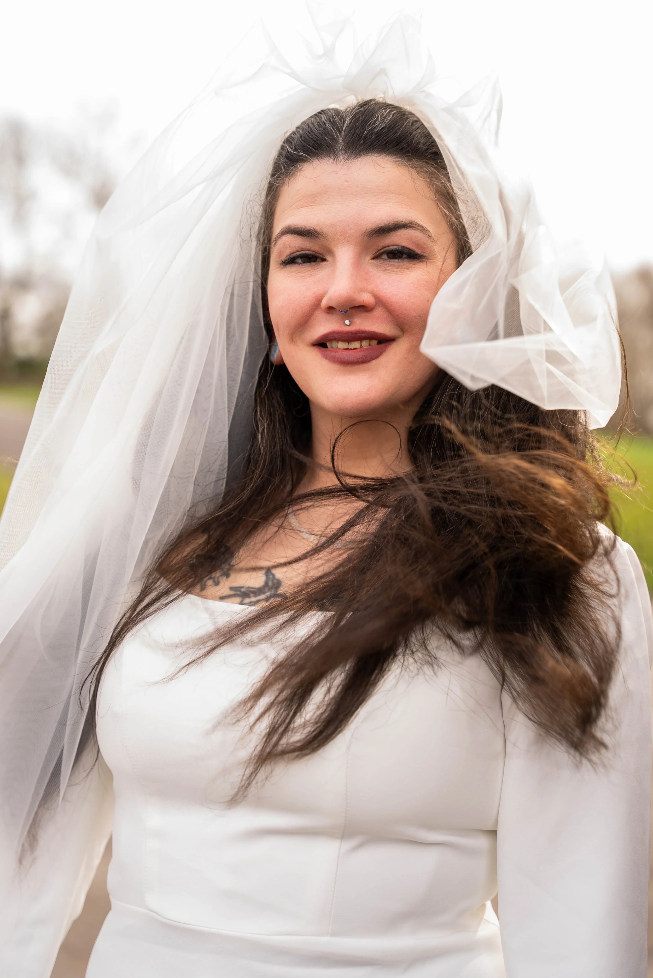 A woman with long dark hair and makeup, wearing a white dress and veil, smiling outdoors with a blurred background.