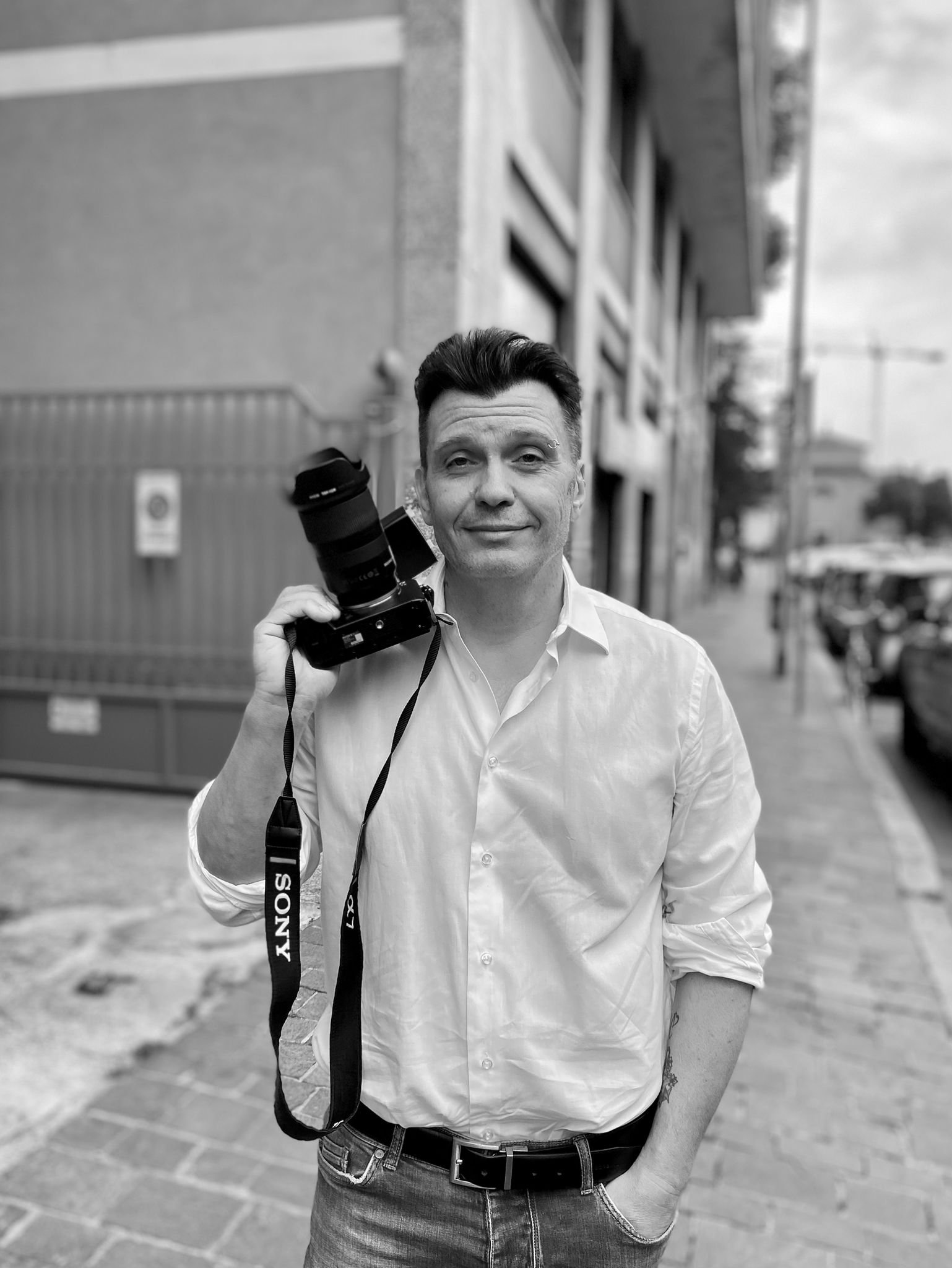 A man in a white shirt holding a camera on his shoulder, standing on an urban sidewalk.