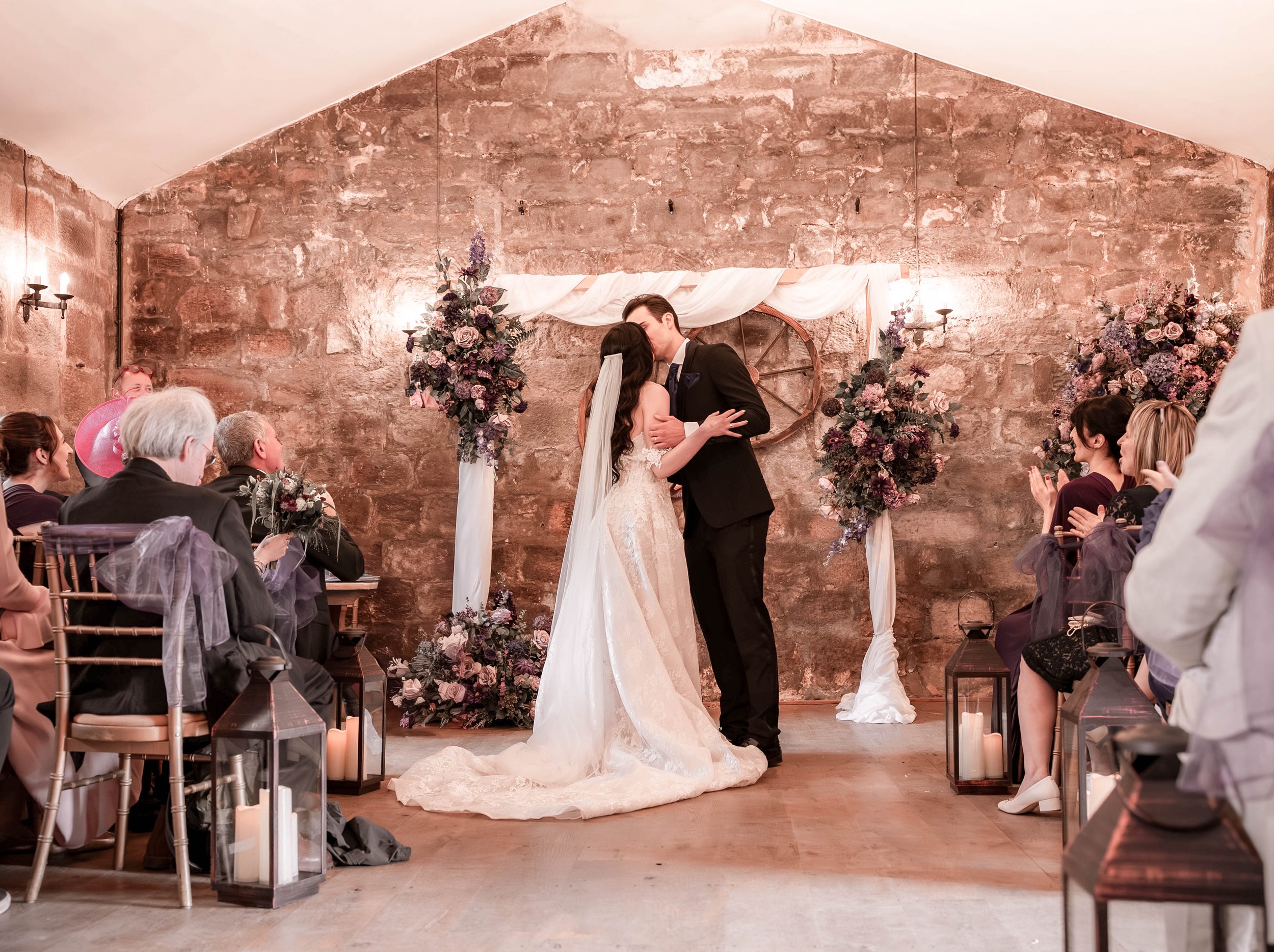 A bride and groom share a kiss during their wedding ceremony in a rustic indoor venue with exposed brick walls, floral decorations, lanterns, and candles, surrounded by seated guests.