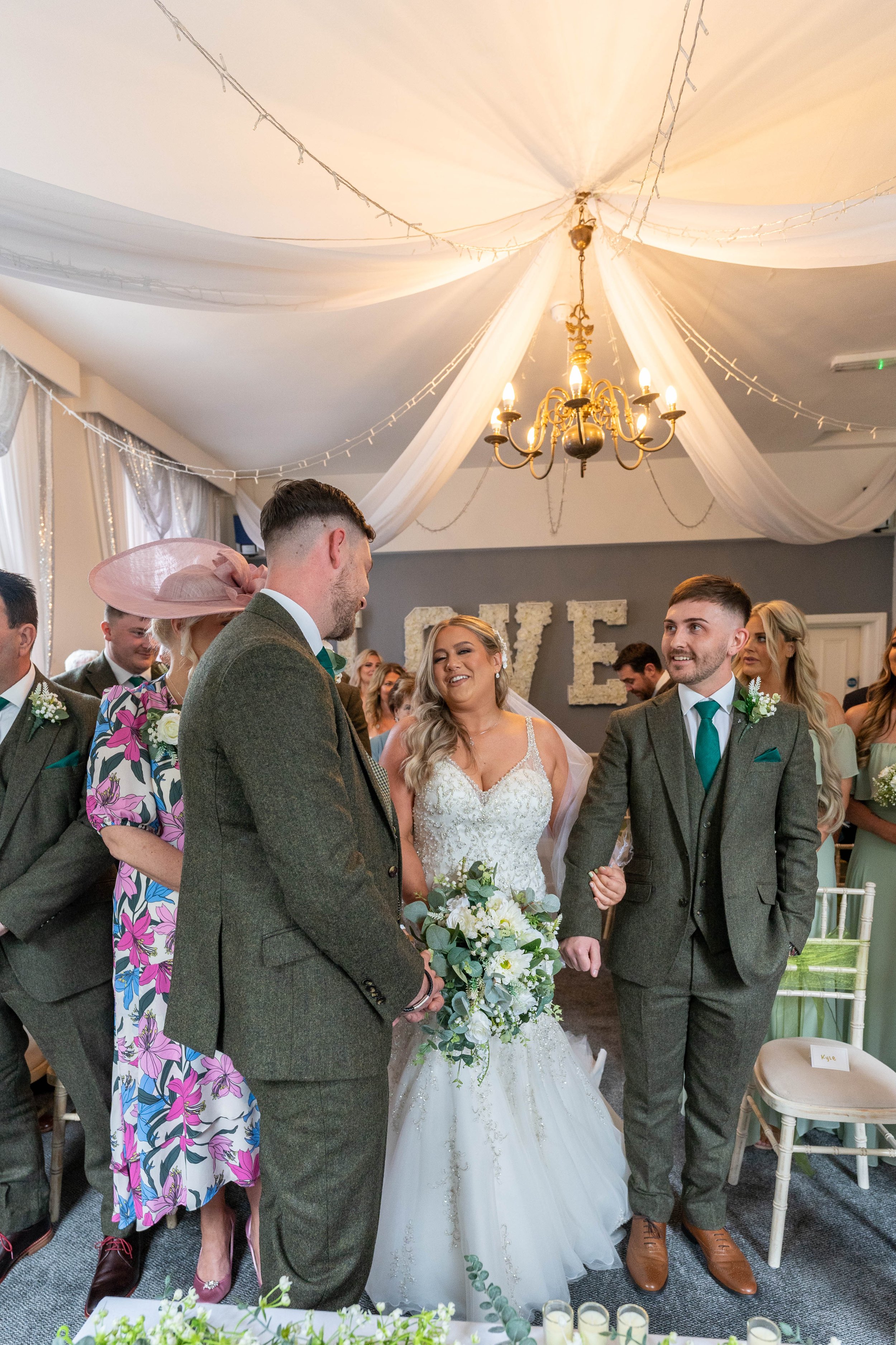Bride and groom standing together, smiling and holding hands during wedding ceremony with guests watching indoors decorated with draped fabric and a chandelier.