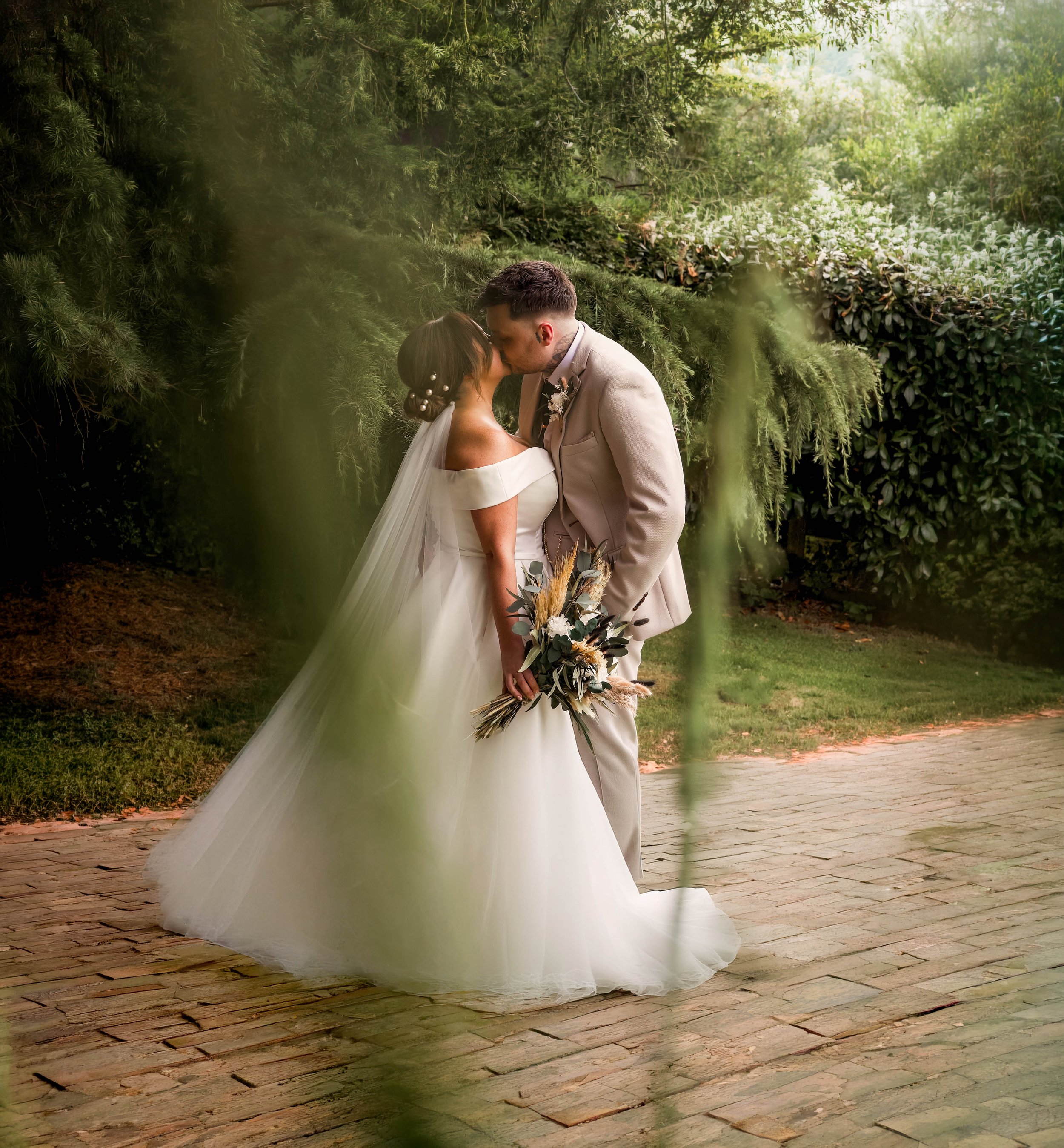 A wedding couple sharing a kiss outdoors, the bride in a white off-shoulder gown holding a bouquet, and the groom in a light-colored suit, surrounded by green foliage.