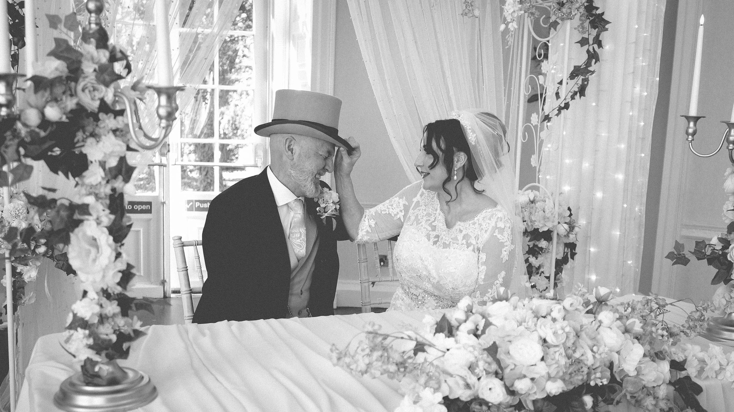 A bride and an older man, possibly her father, share a moment during a wedding celebration, smiling and touching each other's foreheads, surrounded by floral decorations and elegant drapes in a well-lit setting.