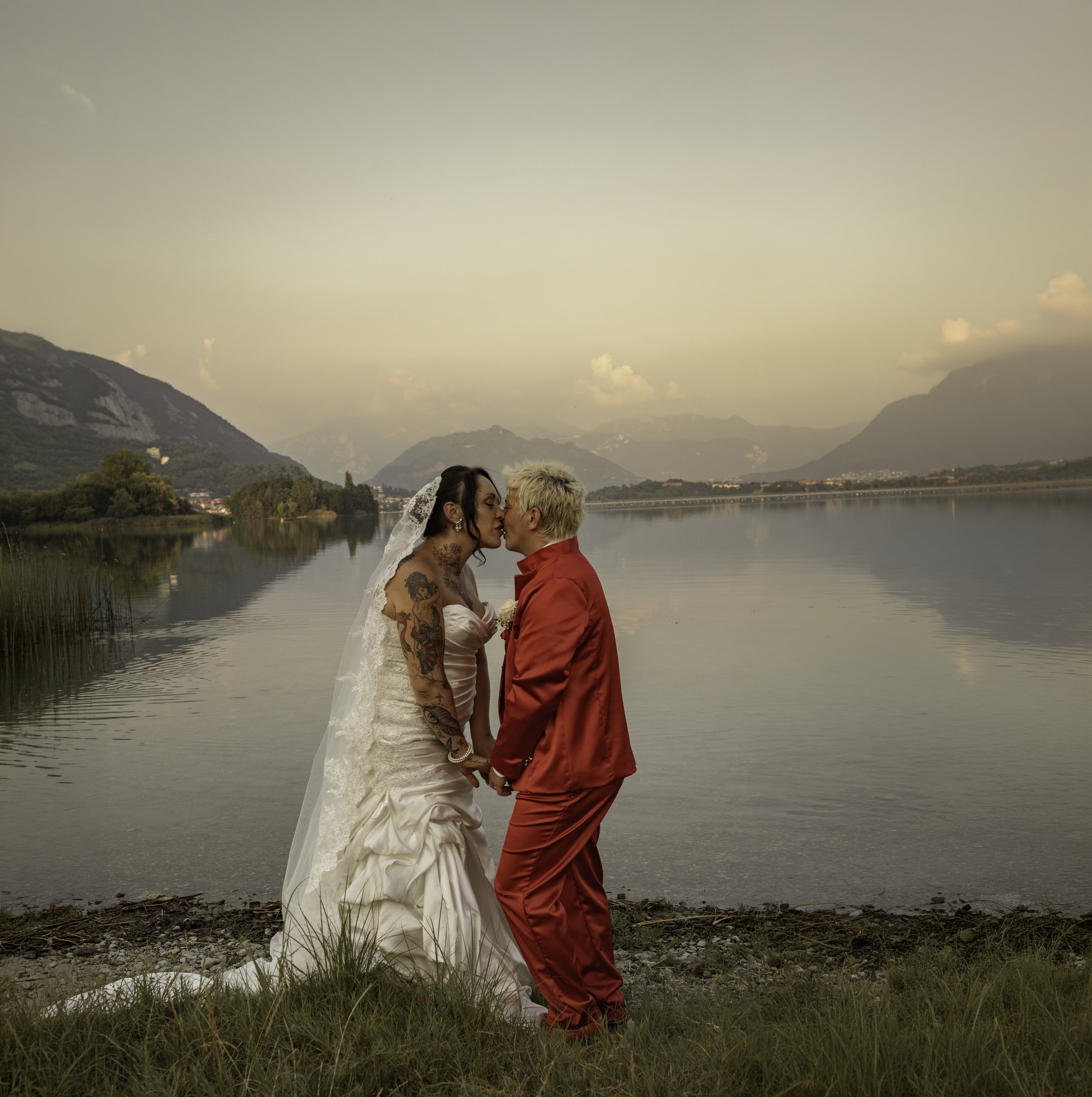 A couple standing by a lake near mountains at sunset, kissing and holding hands, with the woman wearing a wedding dress and the man in a red jacket.