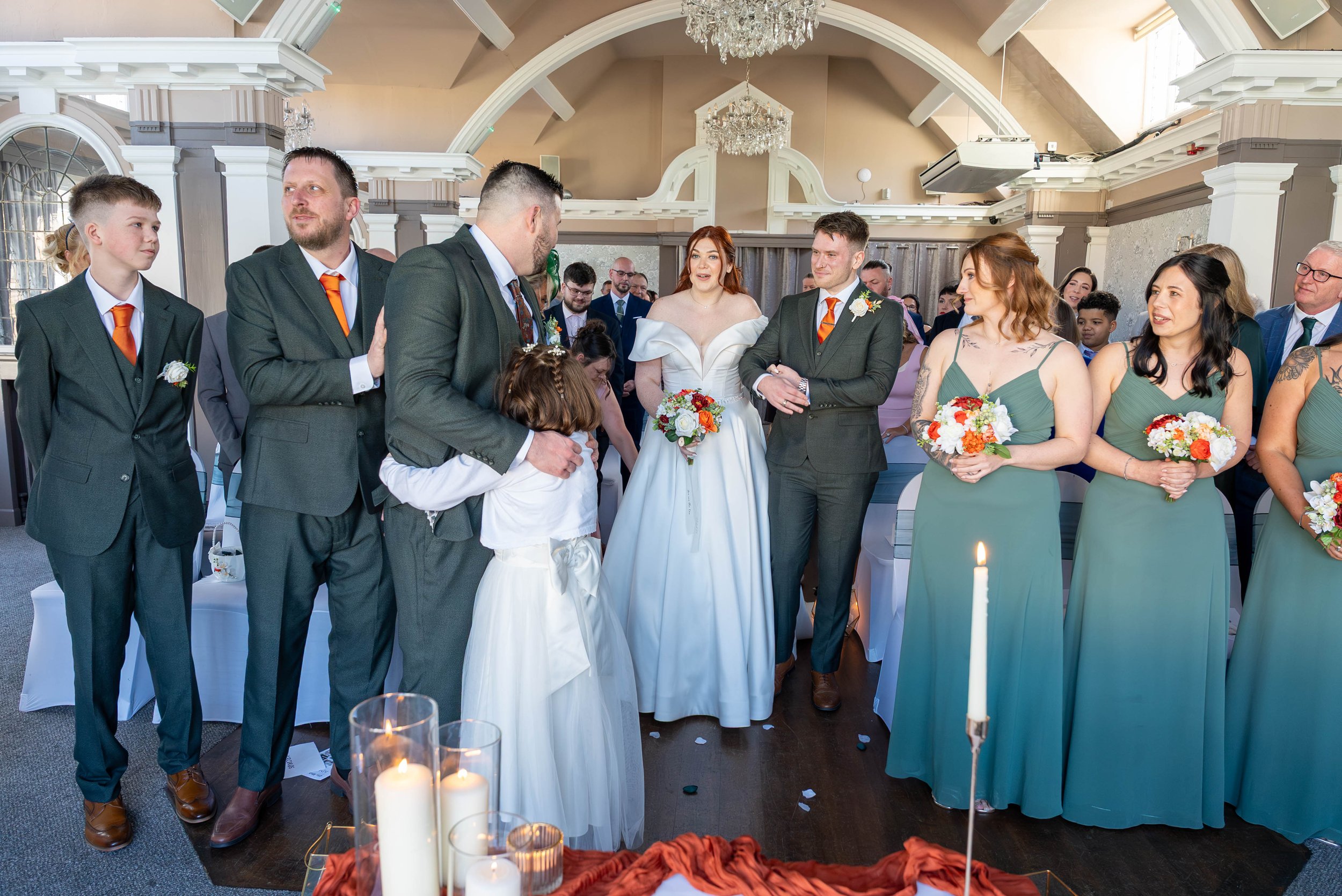 A wedding ceremony with a bride in a white dress holding a bouquet and a groom in a gray suit with an orange tie. They are surrounded by bridesmaids in matching green dresses holding bouquets, groomsmen in gray suits, and family members. The setting 