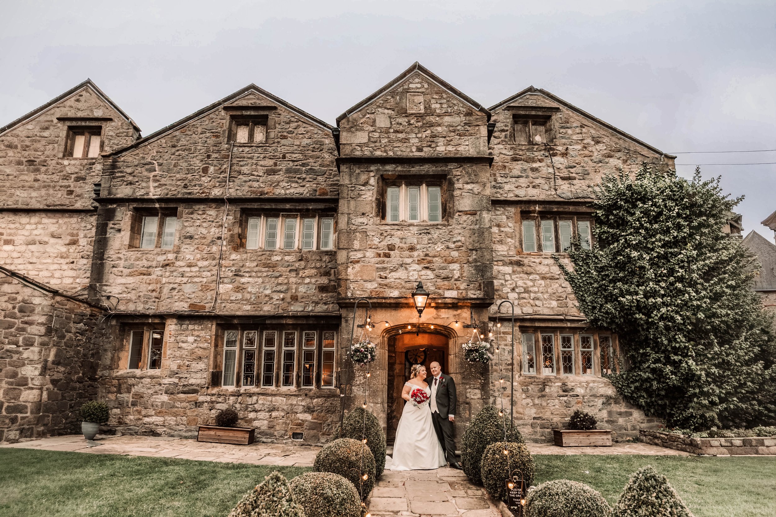 A bride and groom standing together in front of a historic stone building decorated with string lights and hanging flower baskets, during their wedding celebration at dusk.