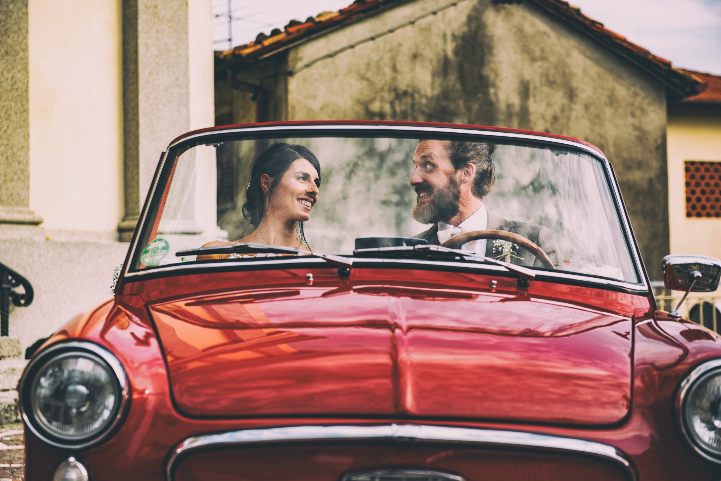 A smiling couple in vintage car, couple looking at each other, woman with dark hair and man with beard, in front of old buildings with tiled roofs.
