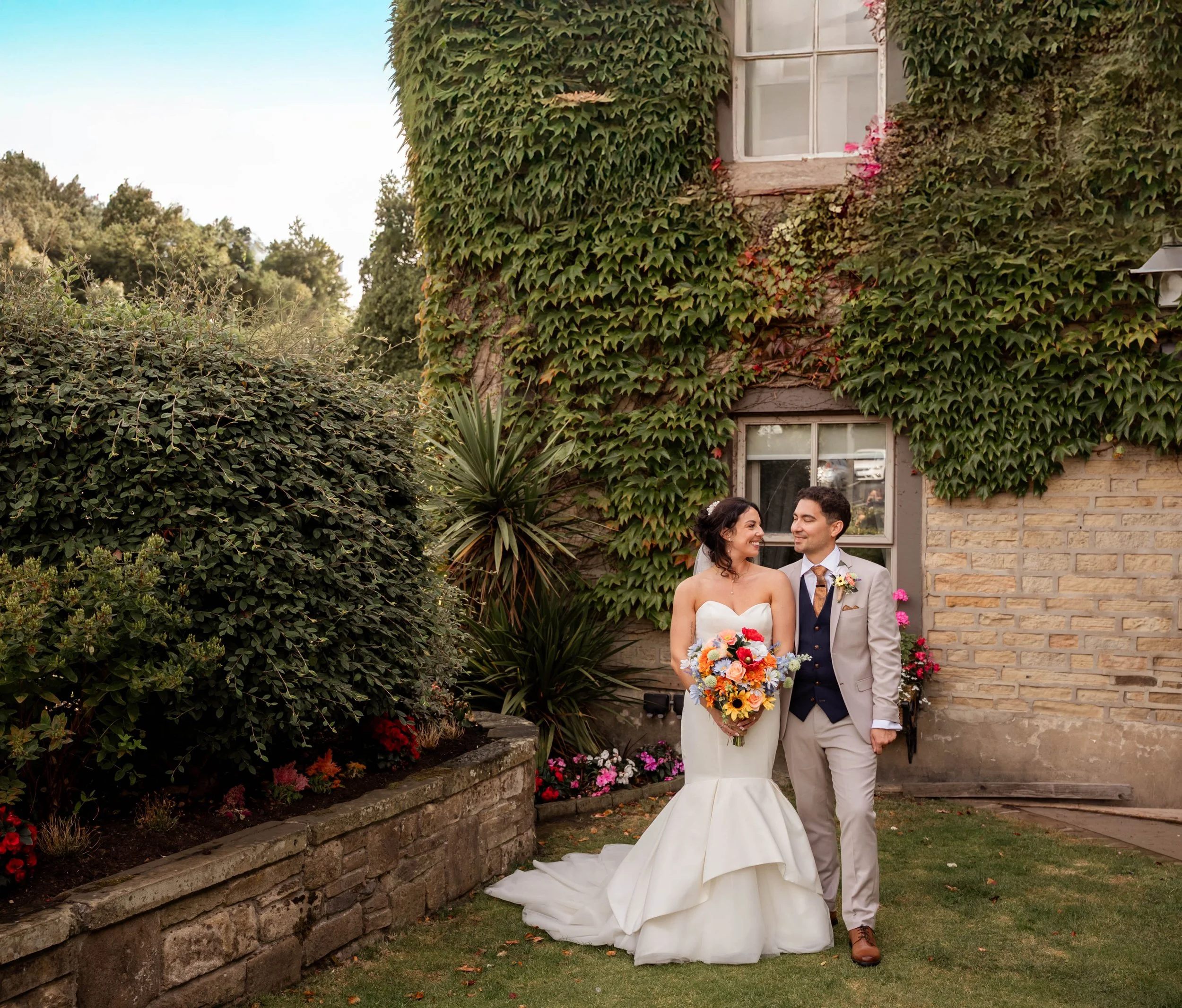 A bride and groom standing outside in front of a house with ivy-covered walls. The bride is holding a colorful bouquet and wearing a strapless white wedding gown. The groom is wearing a light gray suit with a navy vest and is smiling at her.