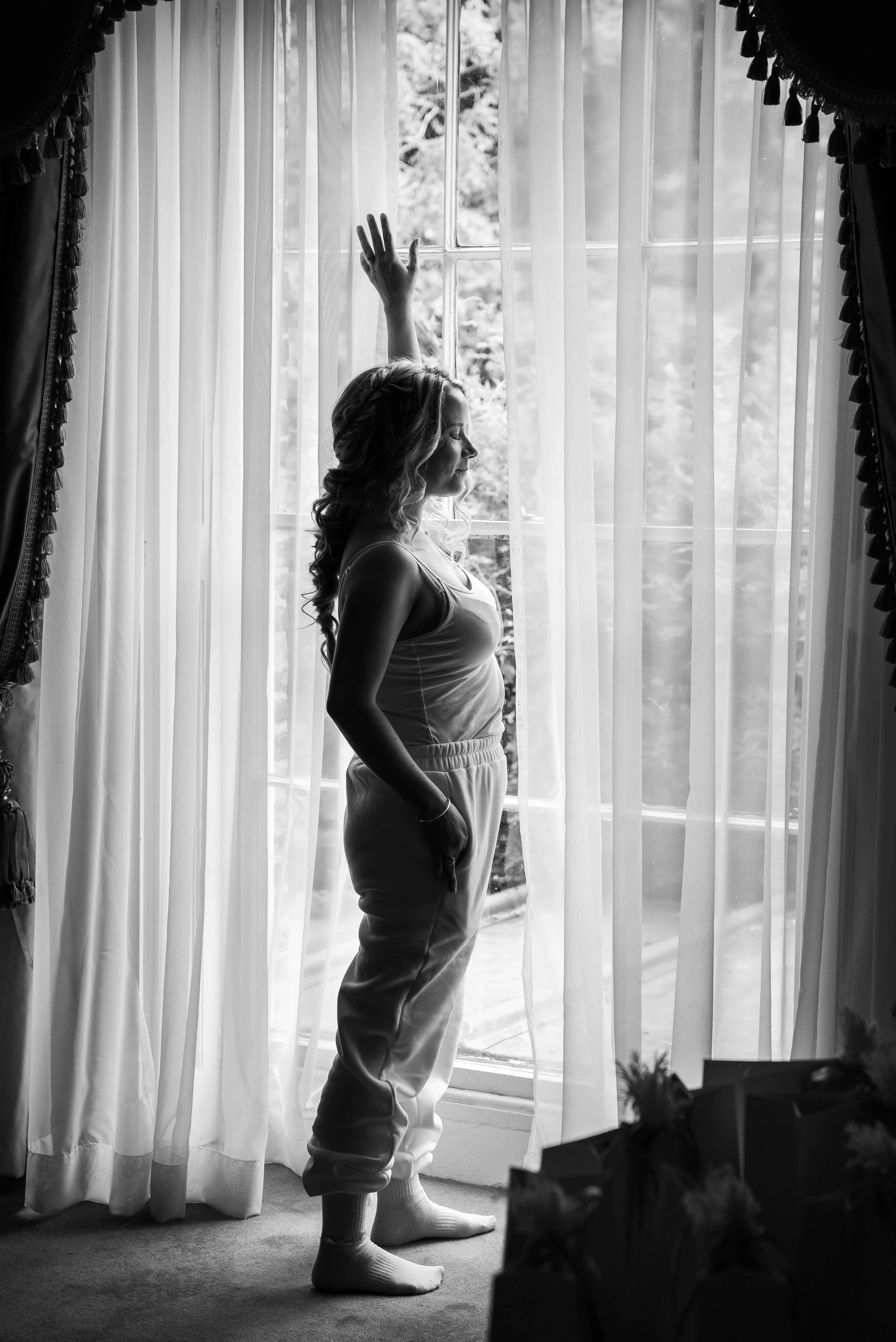 A woman standing by a large window with sheer curtains, her eyes closed and one arm raised, possibly in a moment of reflection or calm, in a room with floral decorations and a soft light.