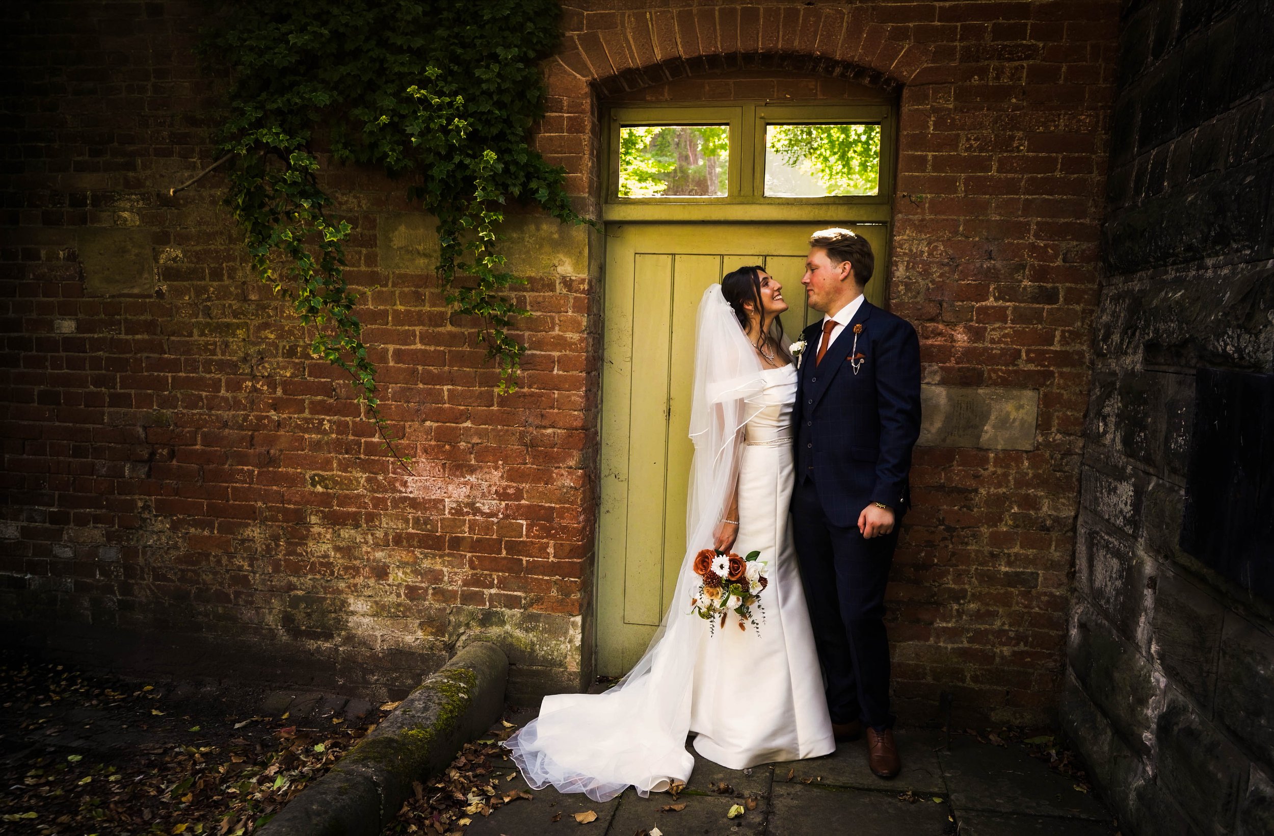 A bride and groom stand close together in front of a green door, smiling and gazing at each other, with a brick wall and ivy in the background.