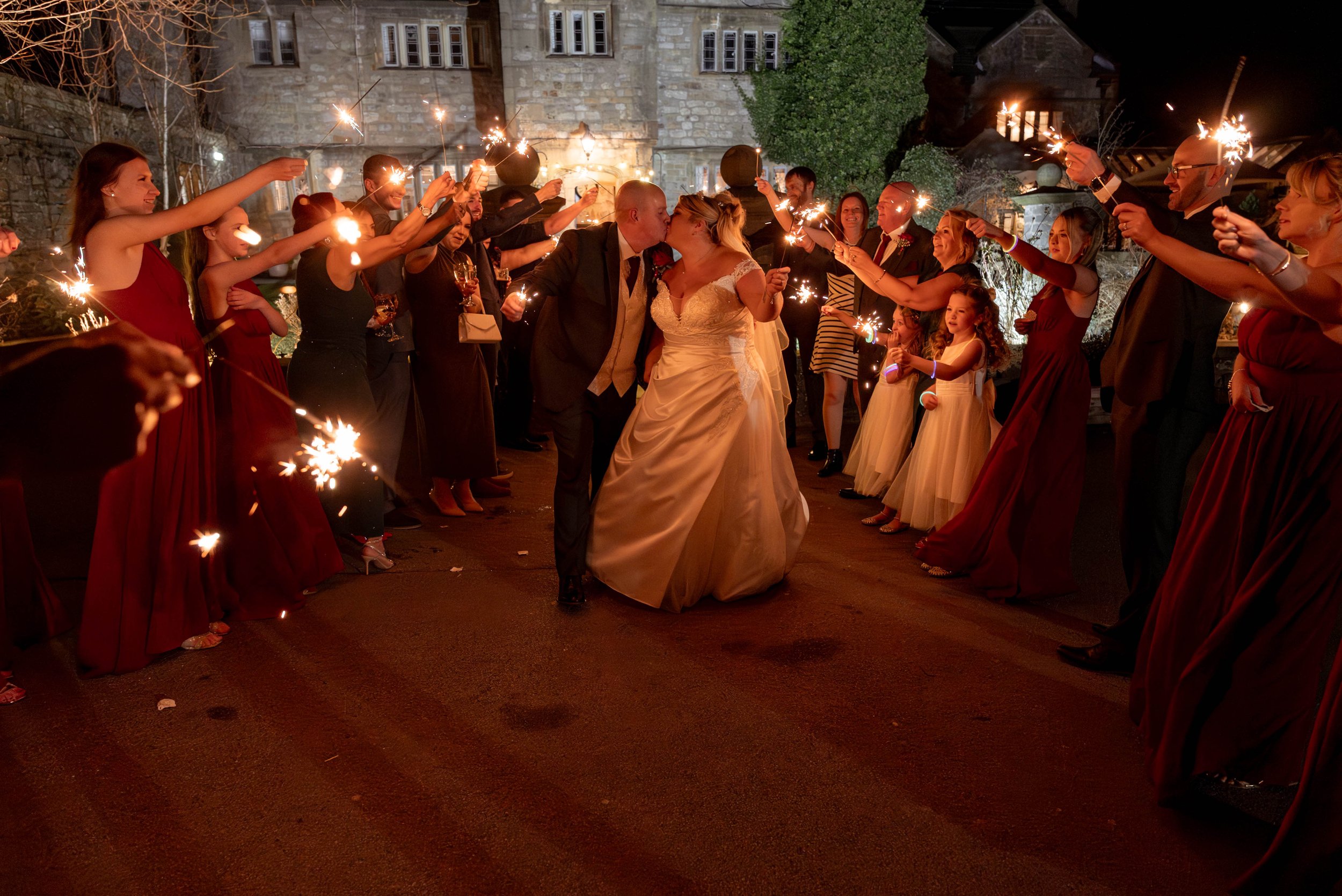 A bride and groom share a kiss surrounded by wedding guests holding sparklers, celebrating at night outside a historic building.