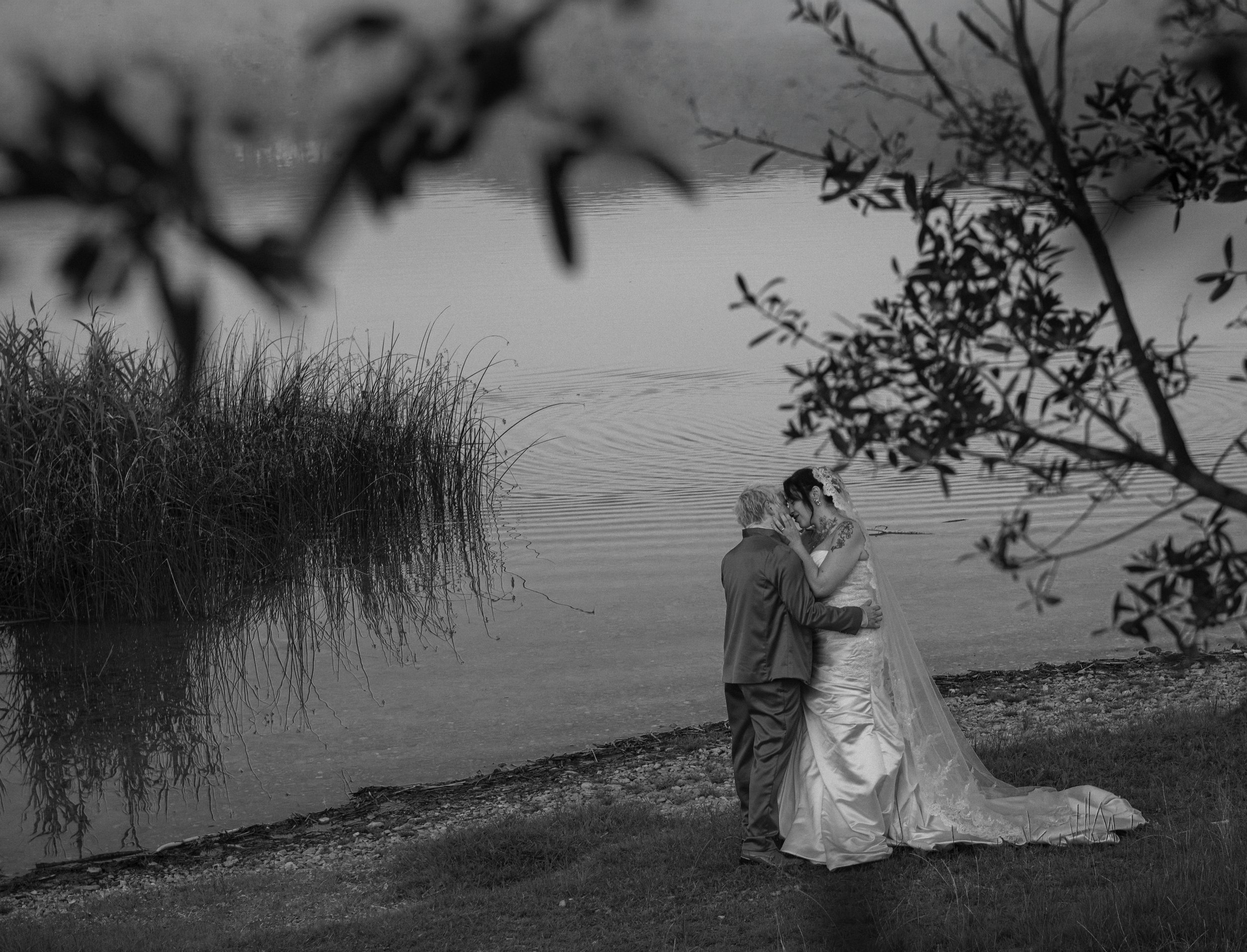 A black and white wedding photograph of a couple kissing by a lakeside with trees and reeds in the background, framed by branches in the foreground.
