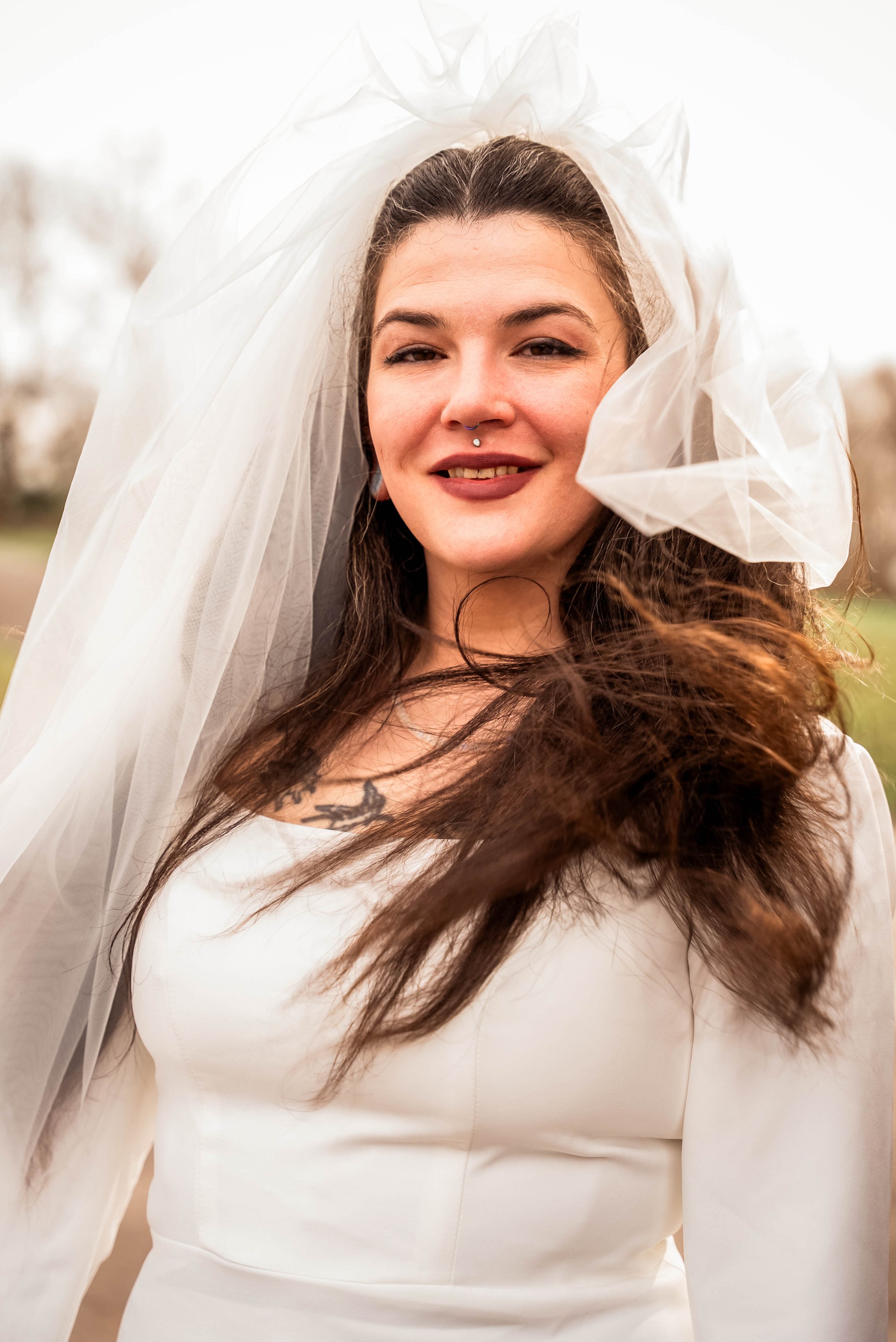 A woman with a septum piercing wearing a white dress and a sheer veil outdoors.