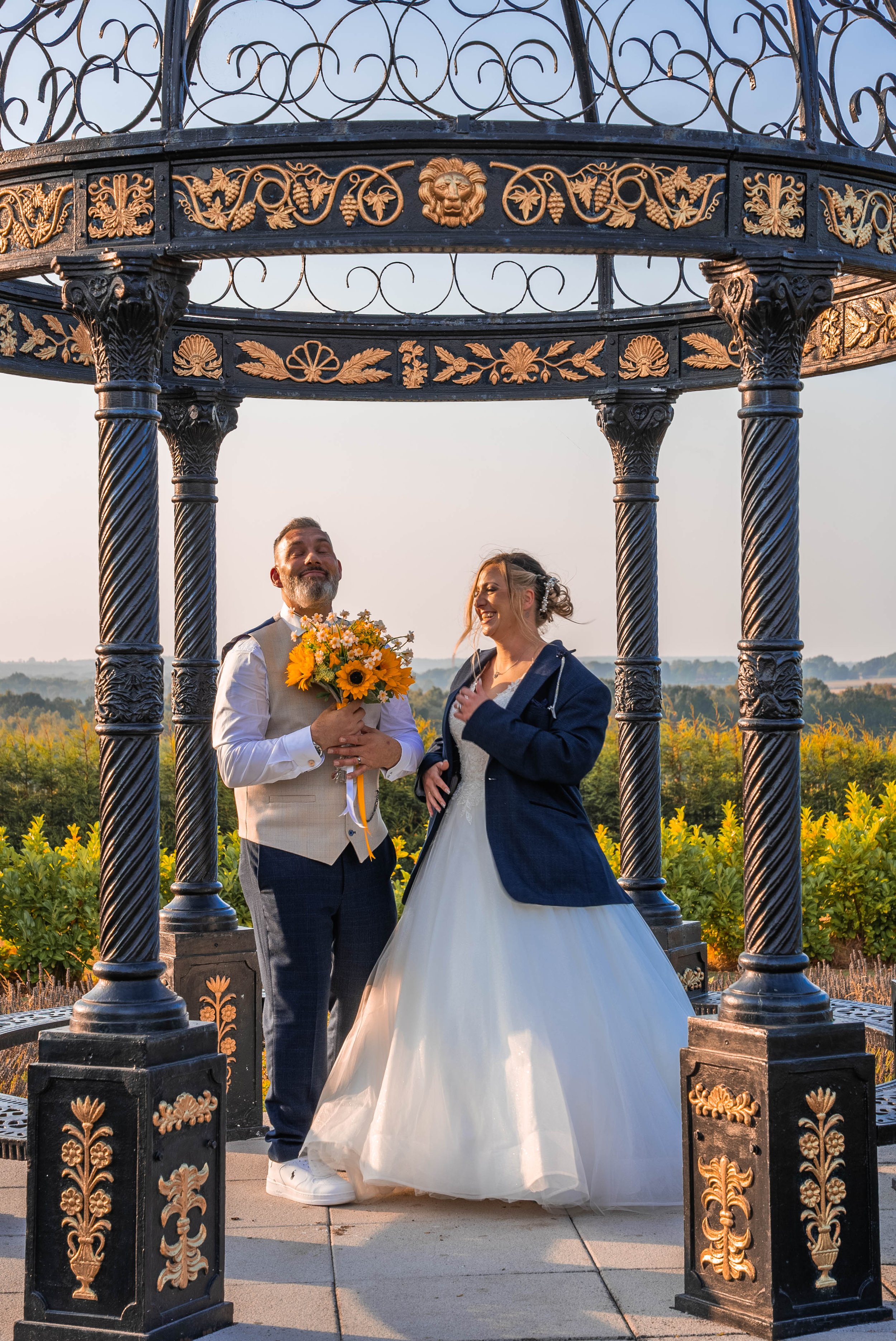 A bride and groom stand beneath an ornate black and gold gazebo, with the groom holding a bouquet of sunflowers and other flowers, during sunset with a landscape of fields and trees in the background.