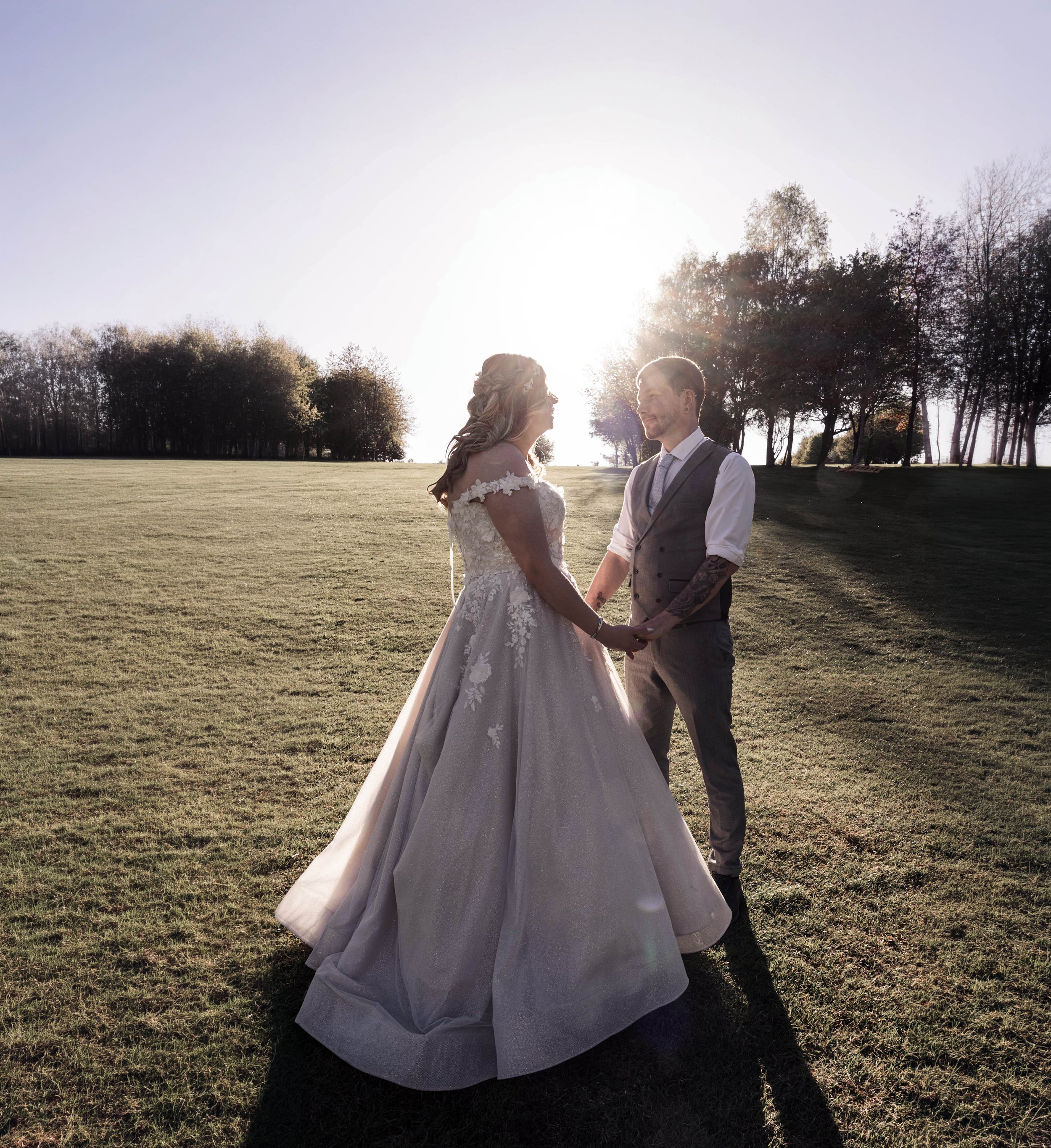 A bride and groom holding hands and gazing at each other outdoors during sunset, with trees in the background.