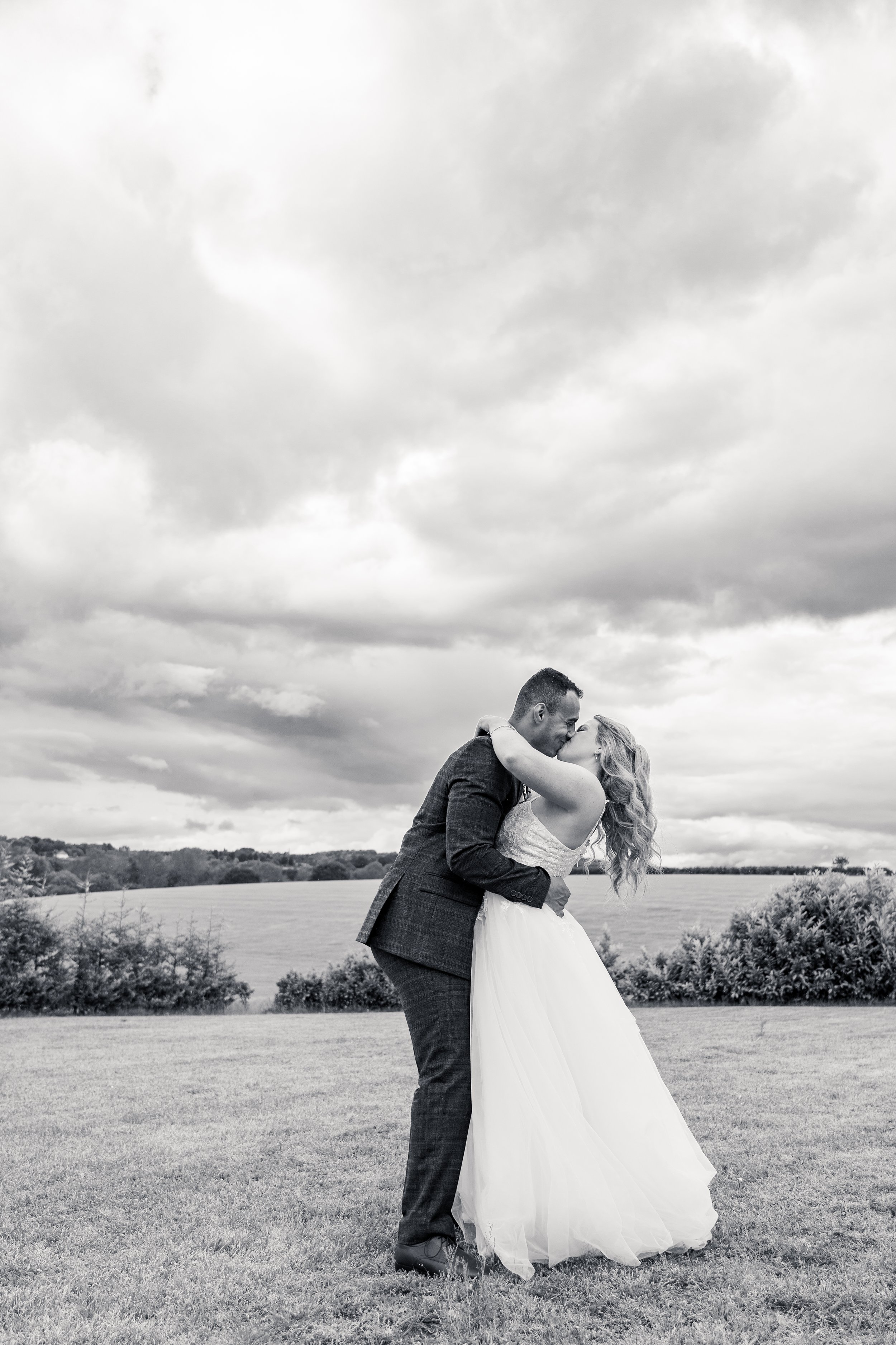 Black and white photo of a couple kissing outdoors, the woman wearing a wedding dress and the man wearing a suit, with a landscape of fields and trees under a cloudy sky in the background.