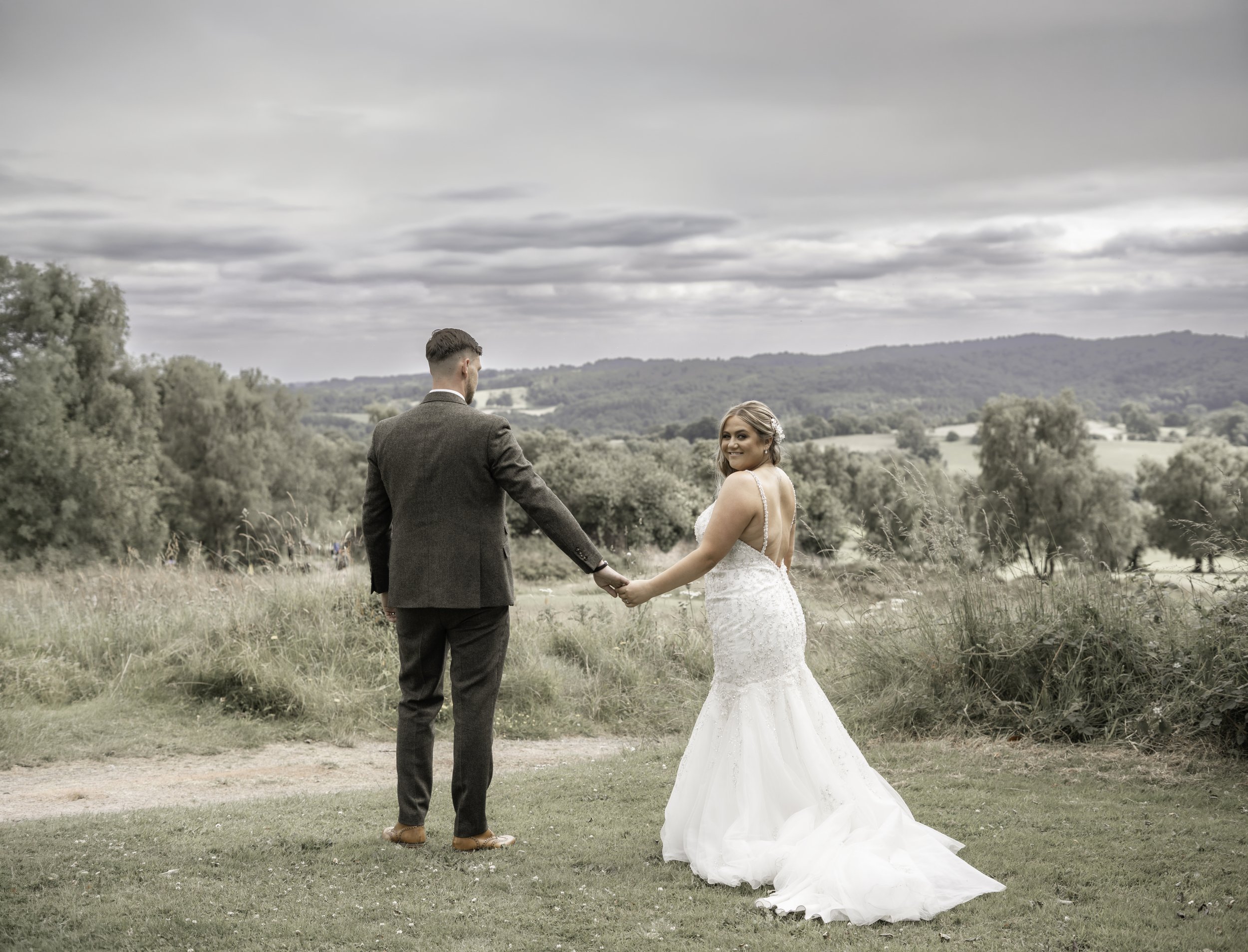 A bride and groom holding hands in an outdoor setting with cloudy sky and rolling hills in the background.