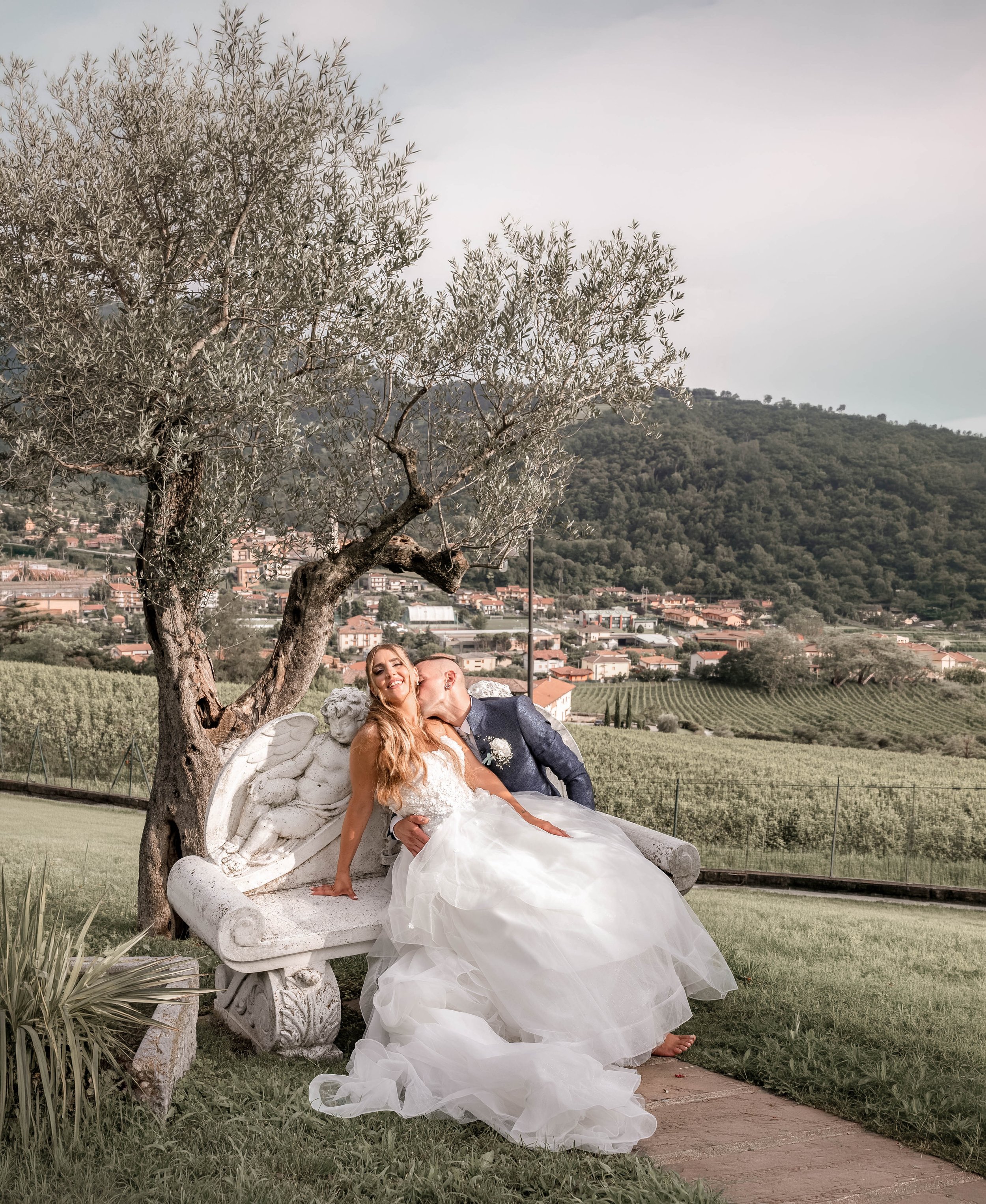 Happy bride and groom sitting on a decorative stone bench outdoors under a tree, with a scenic hilly landscape and rows of grapevines in the background.