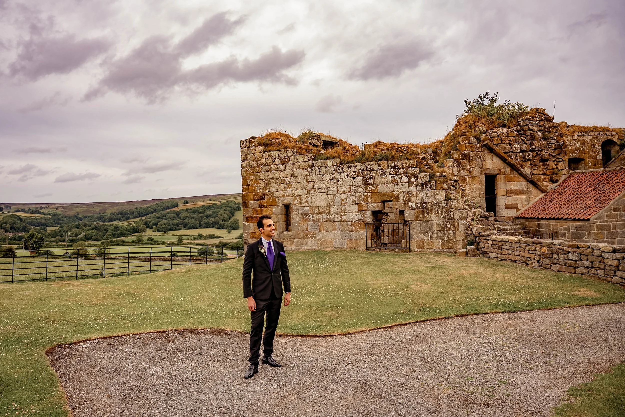 A man in a black suit with a purple tie and pocket square standing outdoors in front of an ancient stone ruin on a cloudy day. The landscape features rolling hills and greenery in the background.