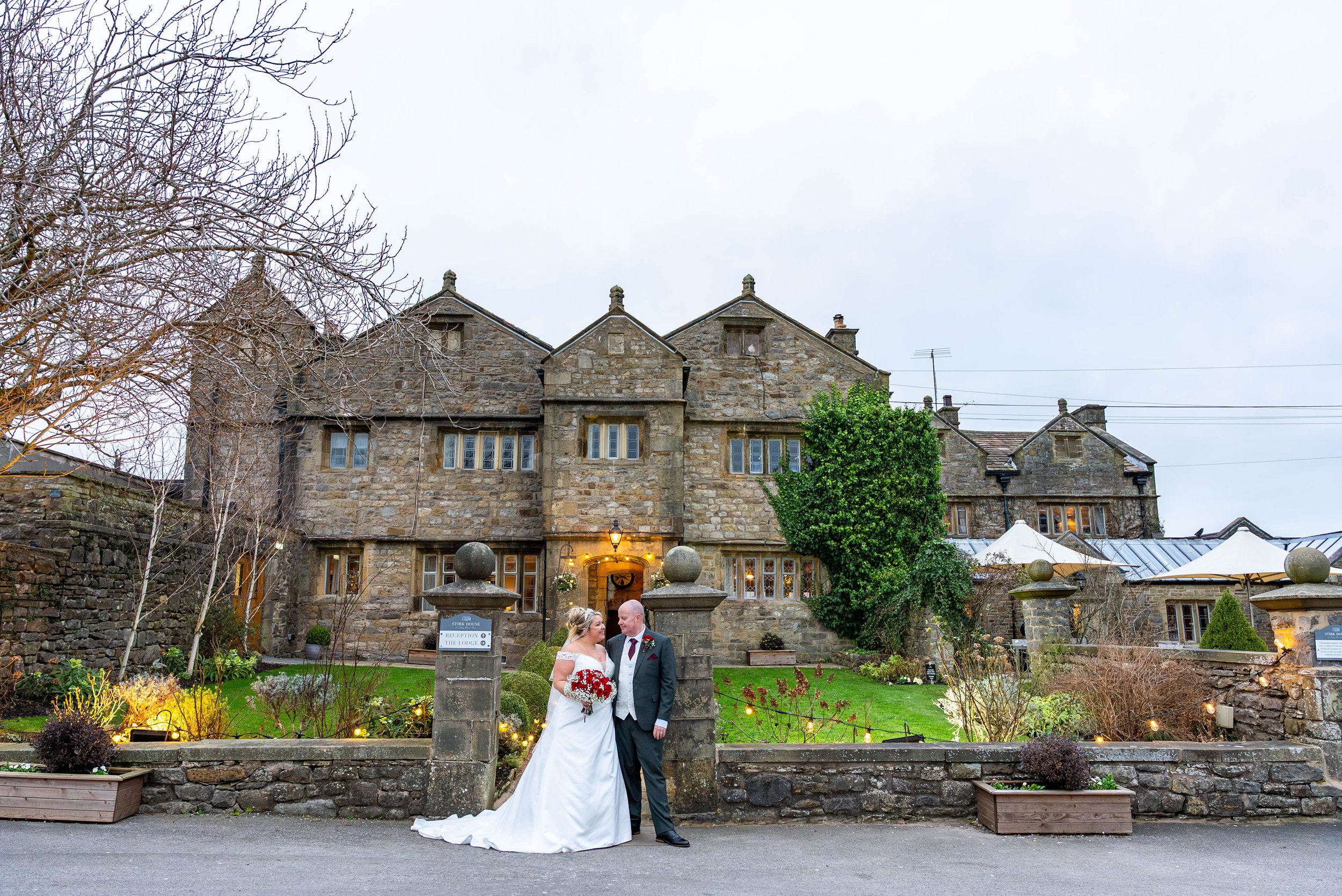 A bride and groom in wedding attire standing in front of a historic stone building with a garden. The bride is holding a bouquet of red and white flowers, and the groom is in a suit. The setting appears to be a wedding ceremony or celebration outdoors in a courtyard with string lights and planters.