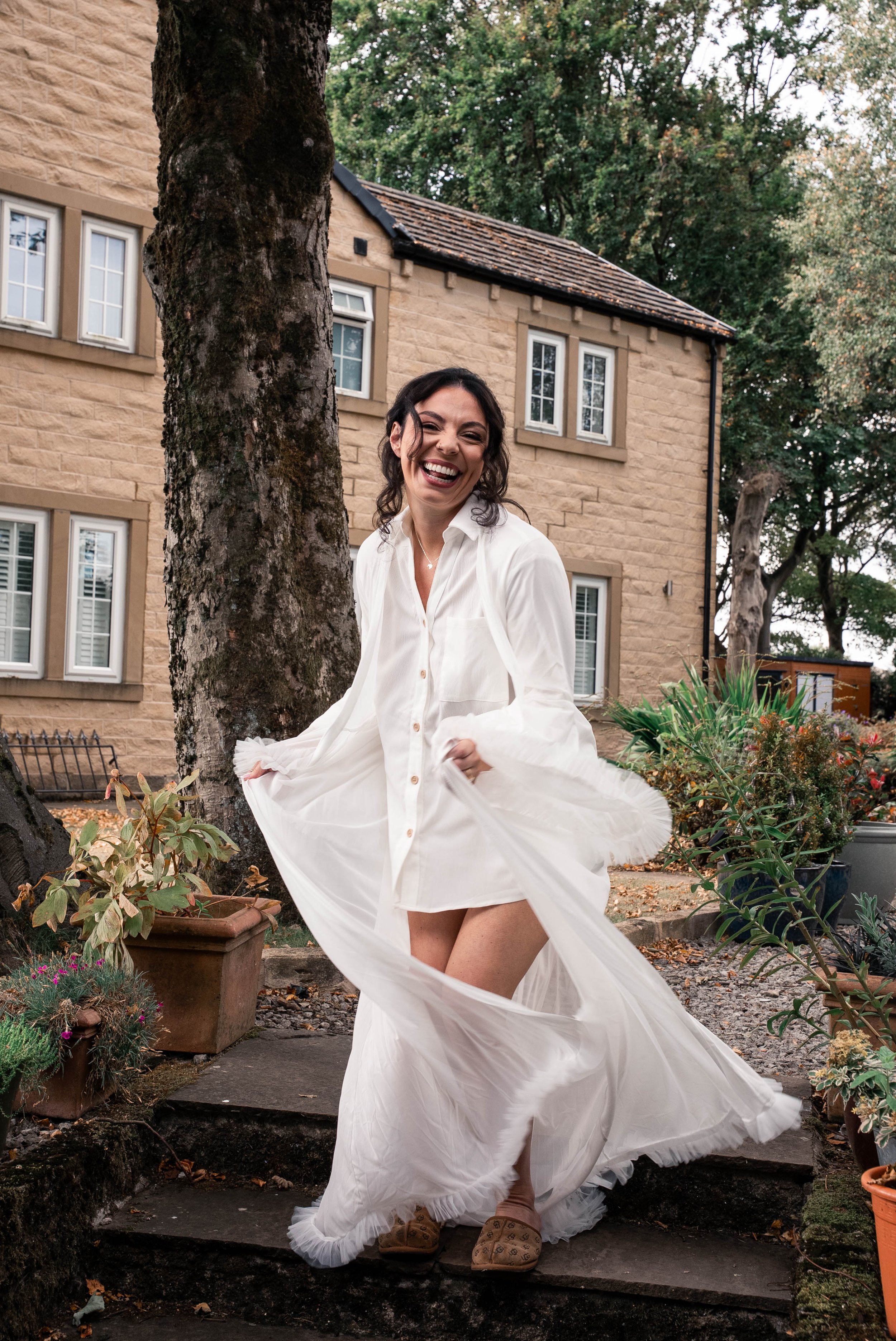 A woman in a white dress smiling and twirling outdoors in front of a tree and a stone house.