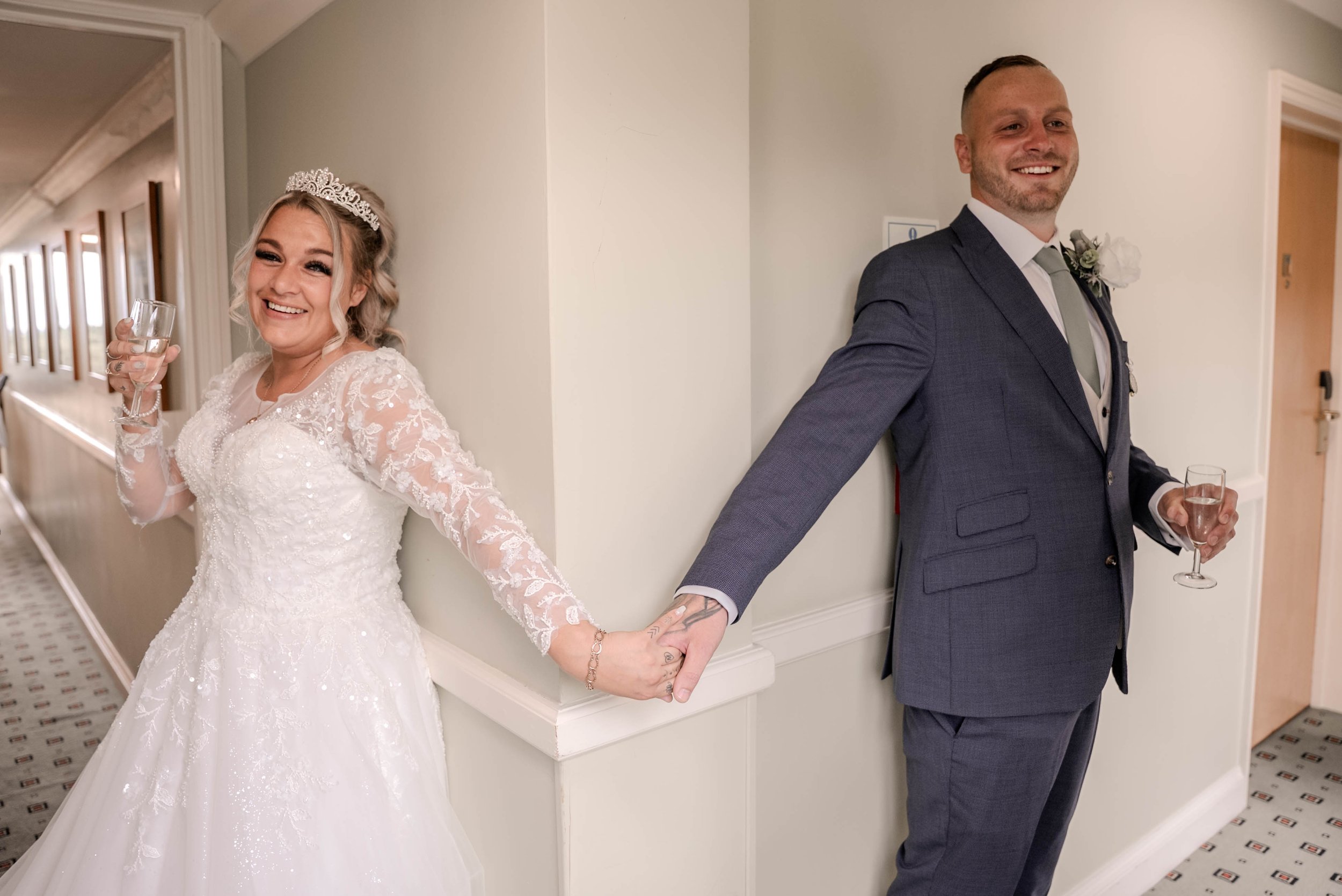 Bride and groom standing back to back, holding hands, each with a glass of champagne, smiling at each other after their wedding.