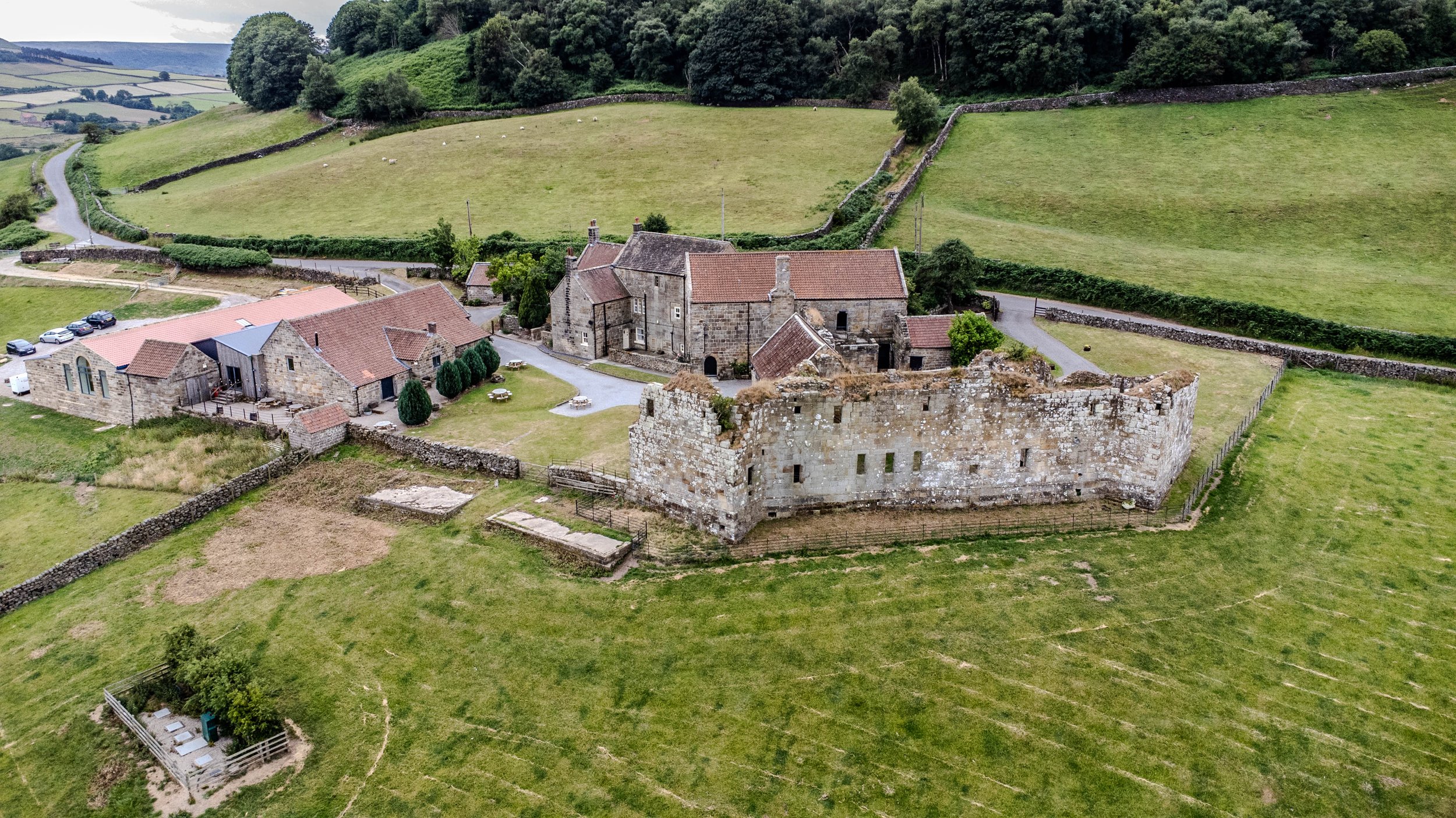 Aerial view of a historic stone castle with surrounding stone buildings, green fields, and hills in the background