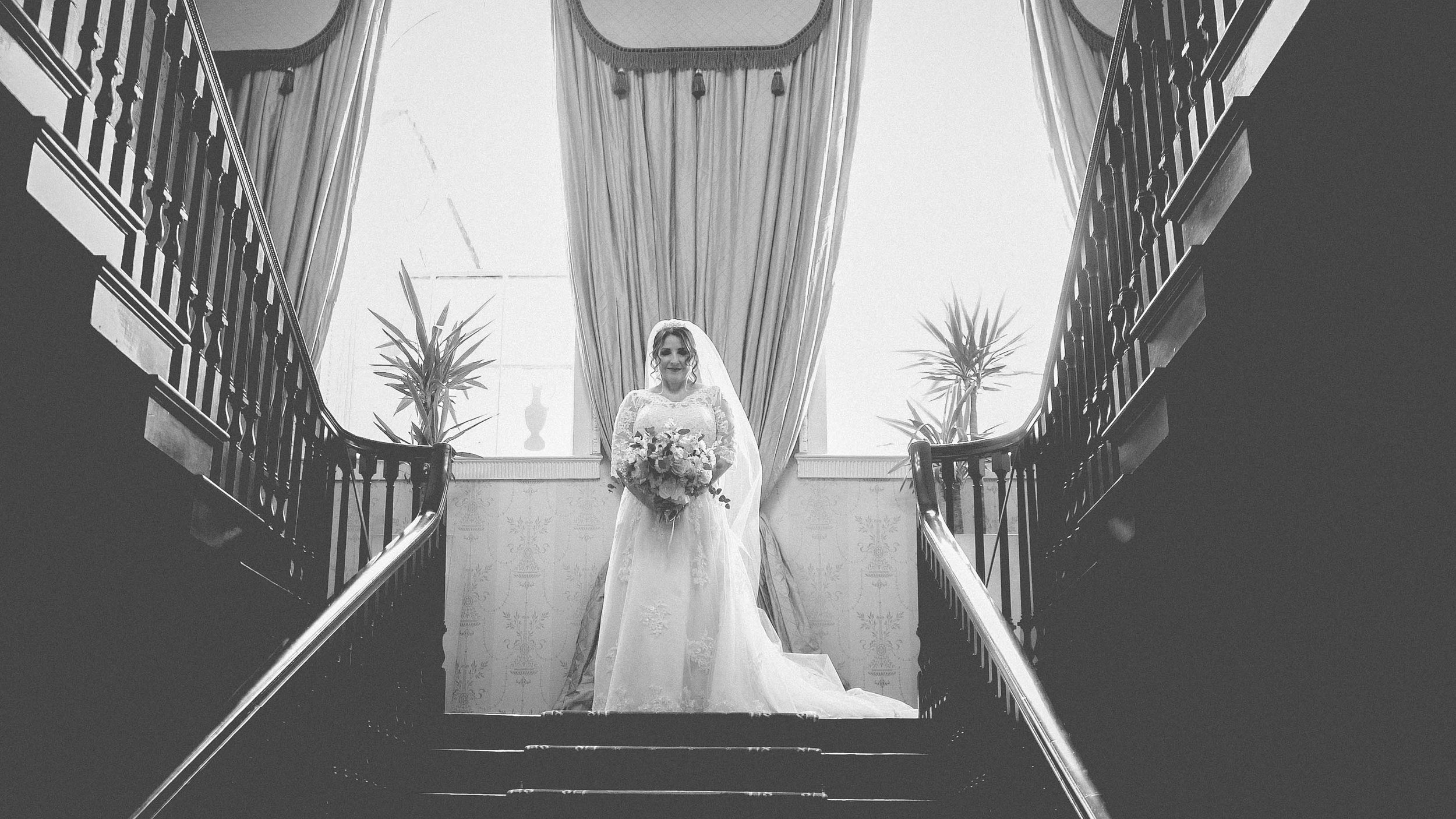 A bride holding a bouquet, standing on a staircase in front of a large window with curtains, inside a grand building.