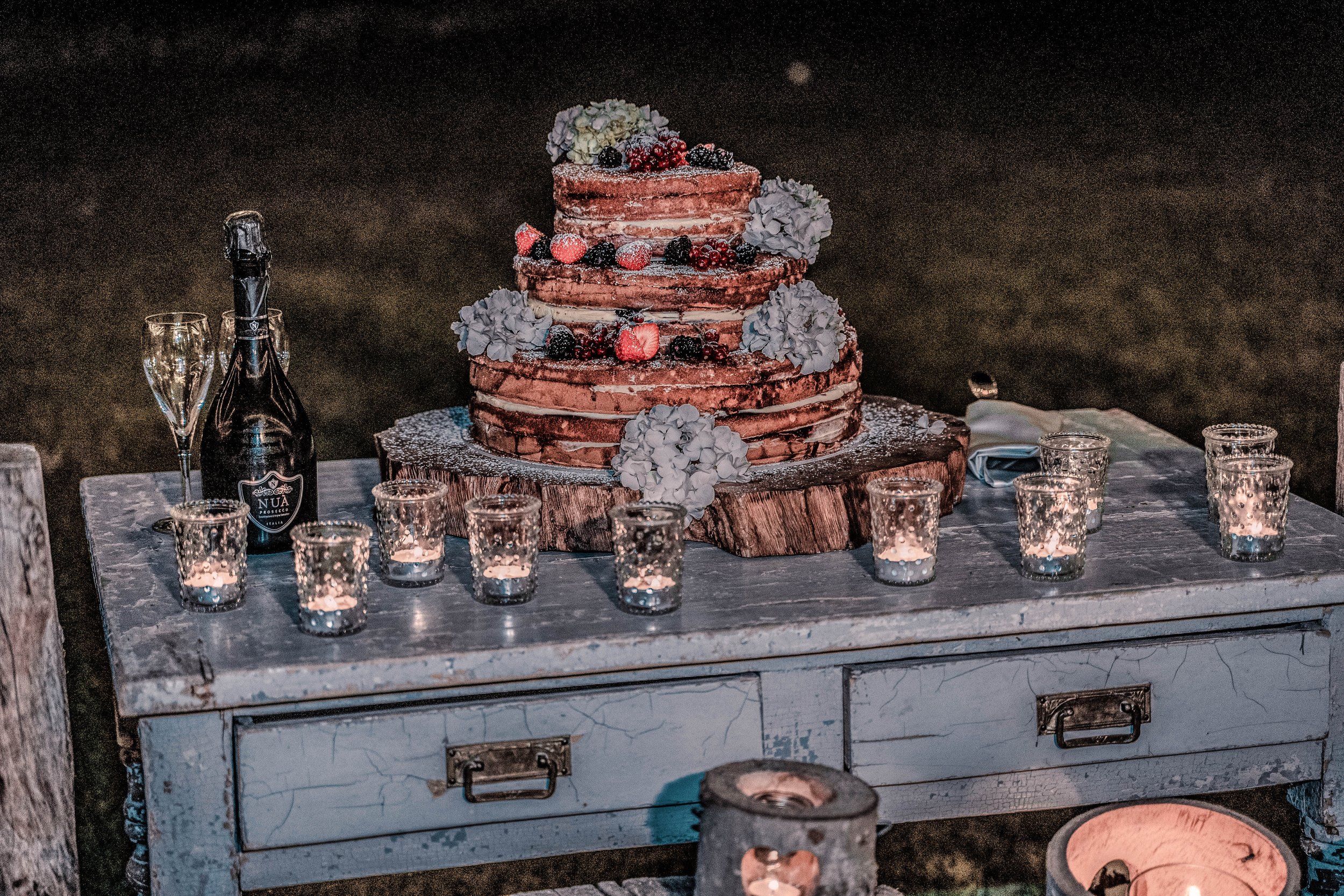 A rustic outdoor celebration table set with a three-tier naked cake decorated with berries and flowers, surrounded by candles in glass holders, with a bottle of champagne and glasses beside it.