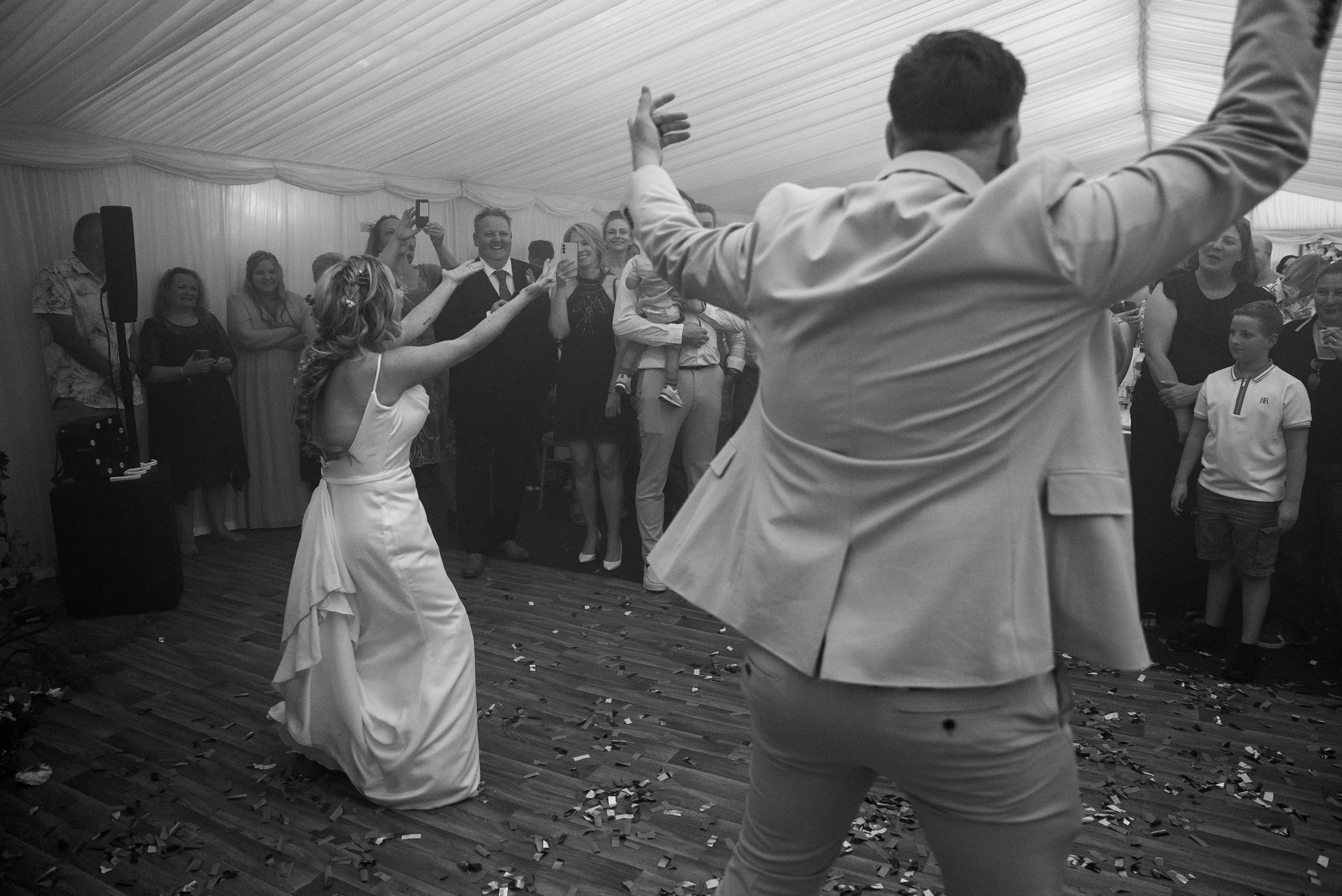 Wedding reception with bride and groom dancing, surrounded by family and friends, under a tent, with confetti on the wooden floor.